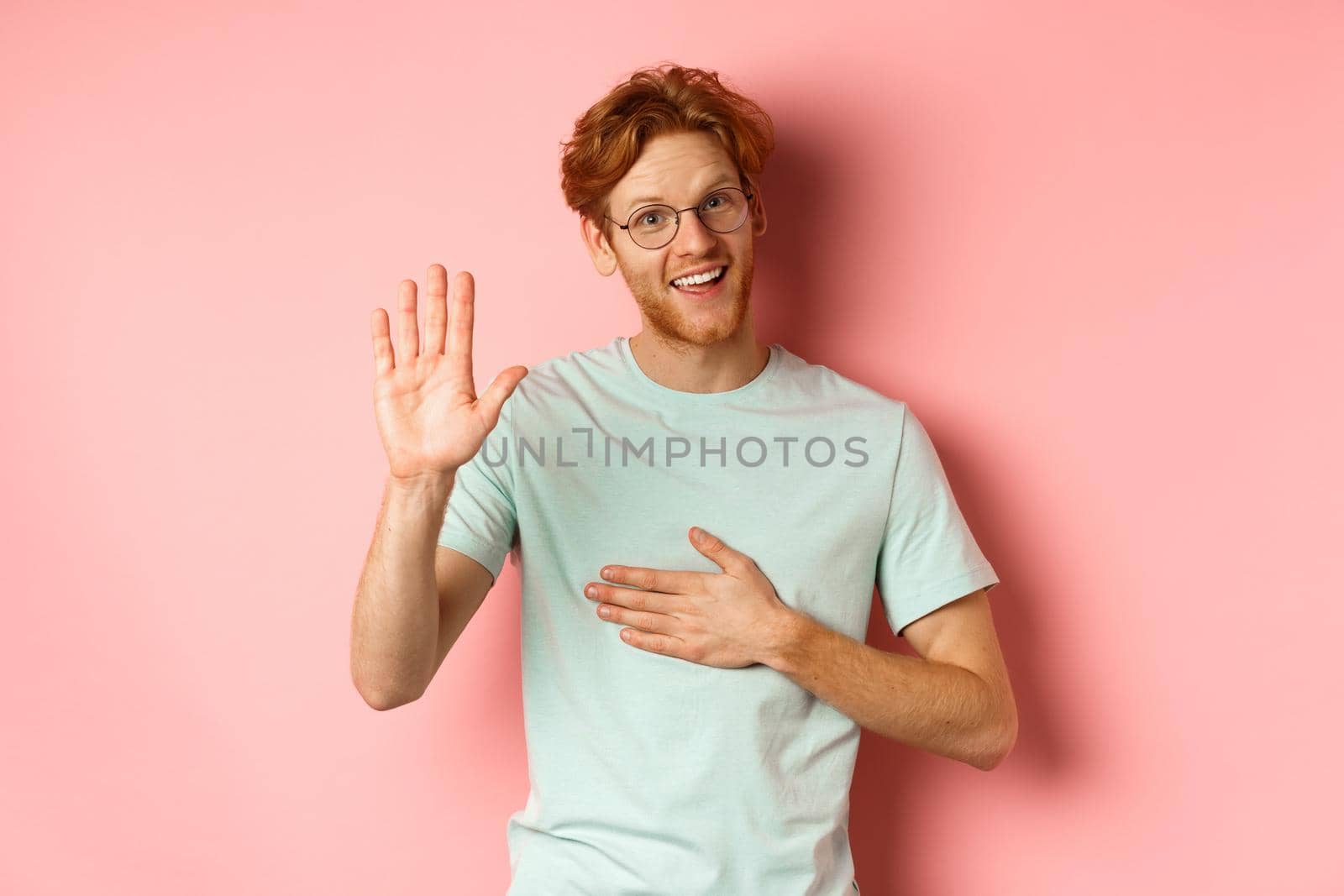 Friendly redhead man being honest, holding hand on heart and arm raised high to swear or make promise, smiling at camera, telling truth over pink background by Benzoix