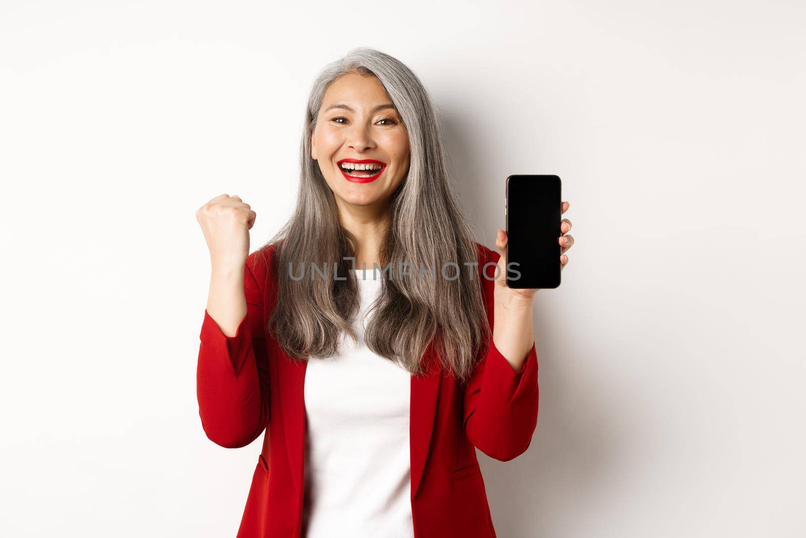 Success. Asian senior woman showing smartphone blank screen and fist pump, winning prize online, standing over white background.
