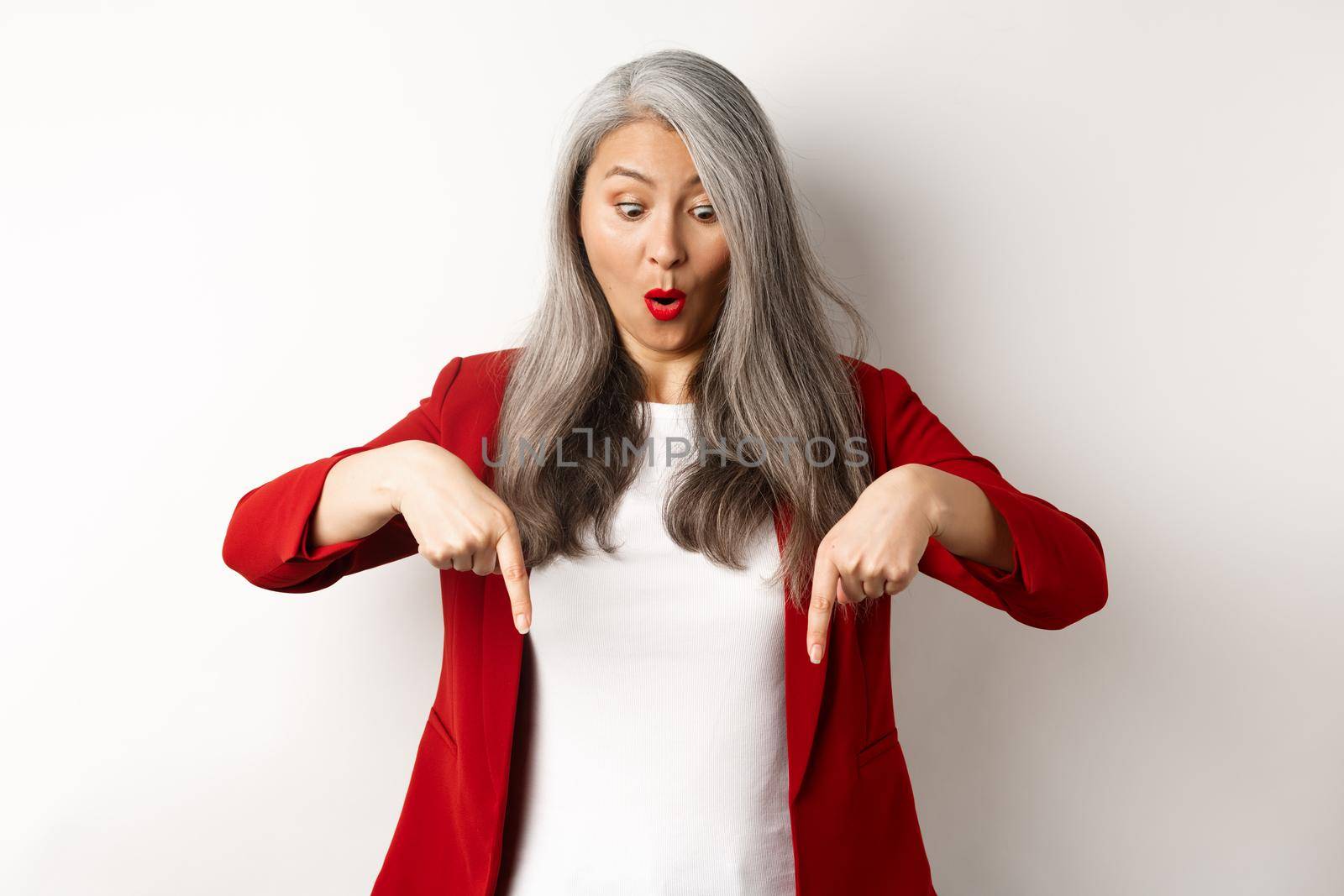 Impressed asian mature woman checking out promo, pointing and looking down with amazed face, standing in blazer over white background by Benzoix