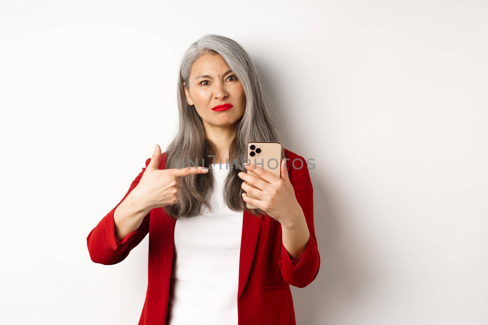 Disappointed mature asian woman complaining, pointing finger at smartphone and looking displeased, standing over white background.