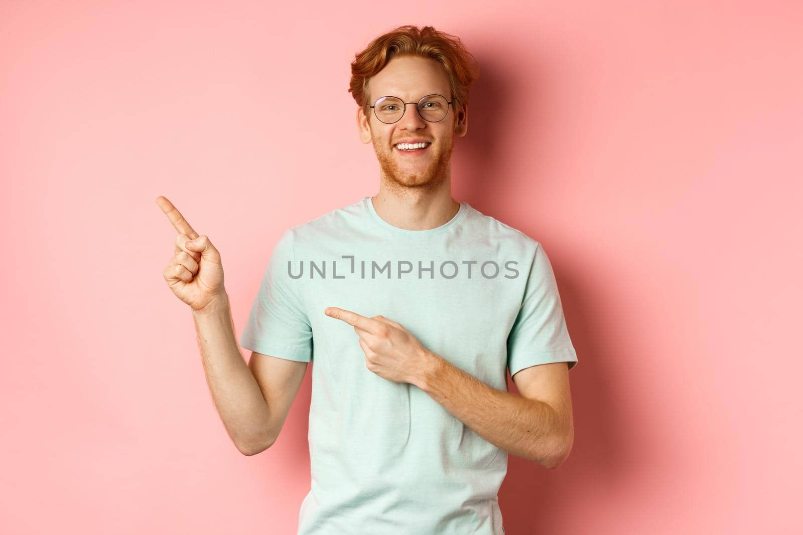 Portrait of cheerful young man with red hair, wearing glasses, pointing fingers at upper left corner and smiling, standing over pink background.