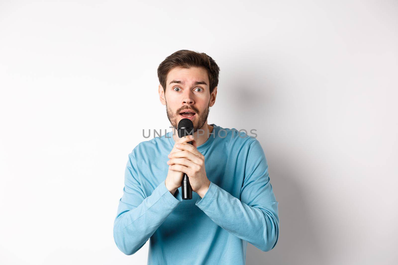 Confused young man looking nervously at camera while singing karaoke, holding microphone, standing over white background.
