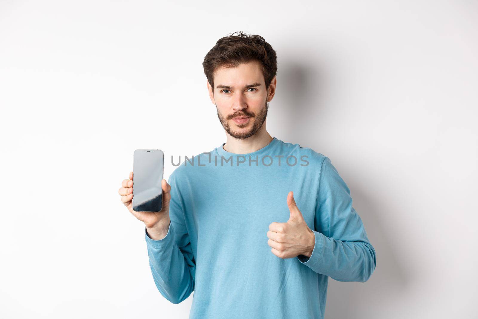 Young man showing empty smartphone screen and thumb up, recommend mobile application or online shopping offer, standing over white background by Benzoix