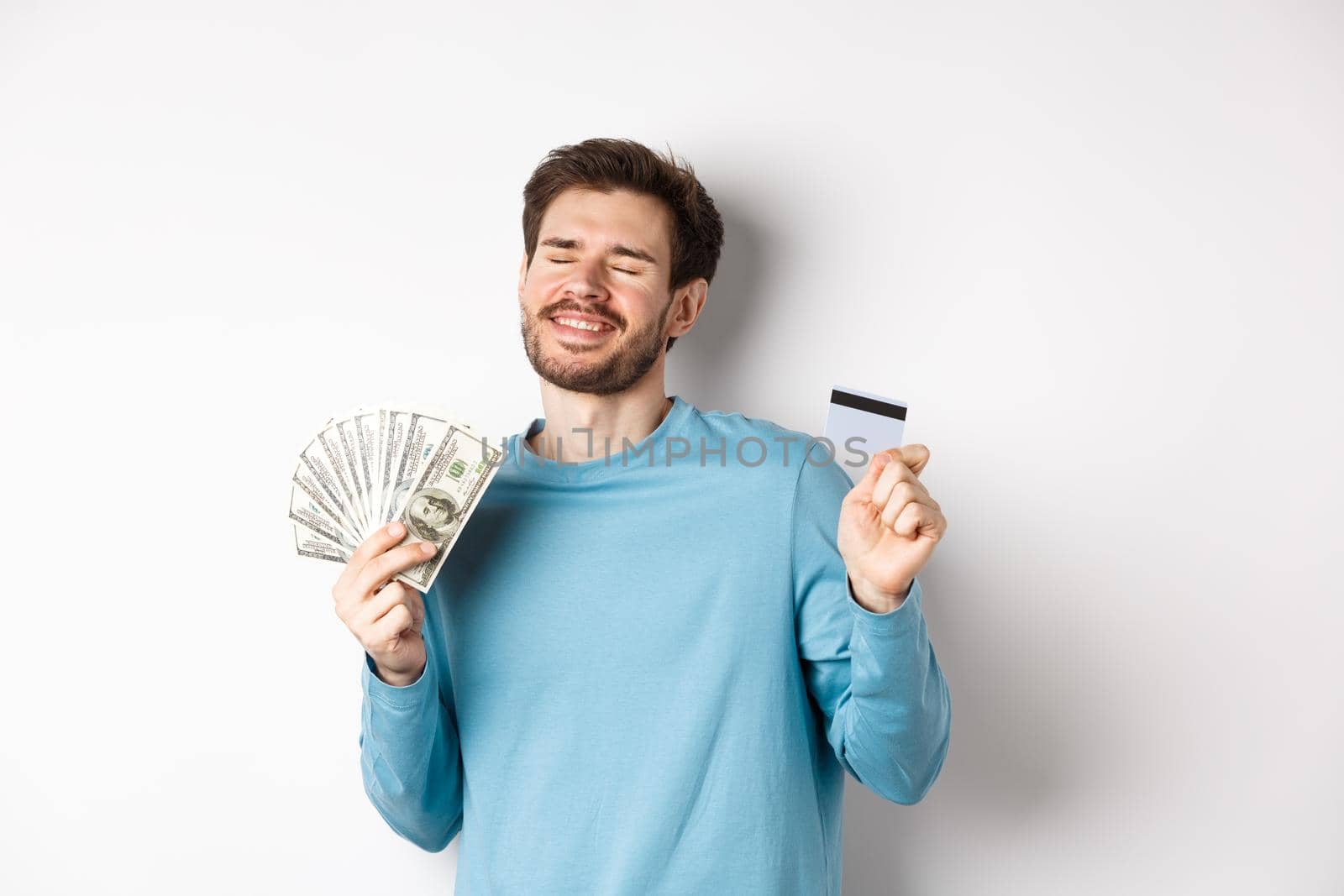 Cheerful guy celebrating salary, dancing with plastic credit card and money, smiling satisfied, standing over white background.