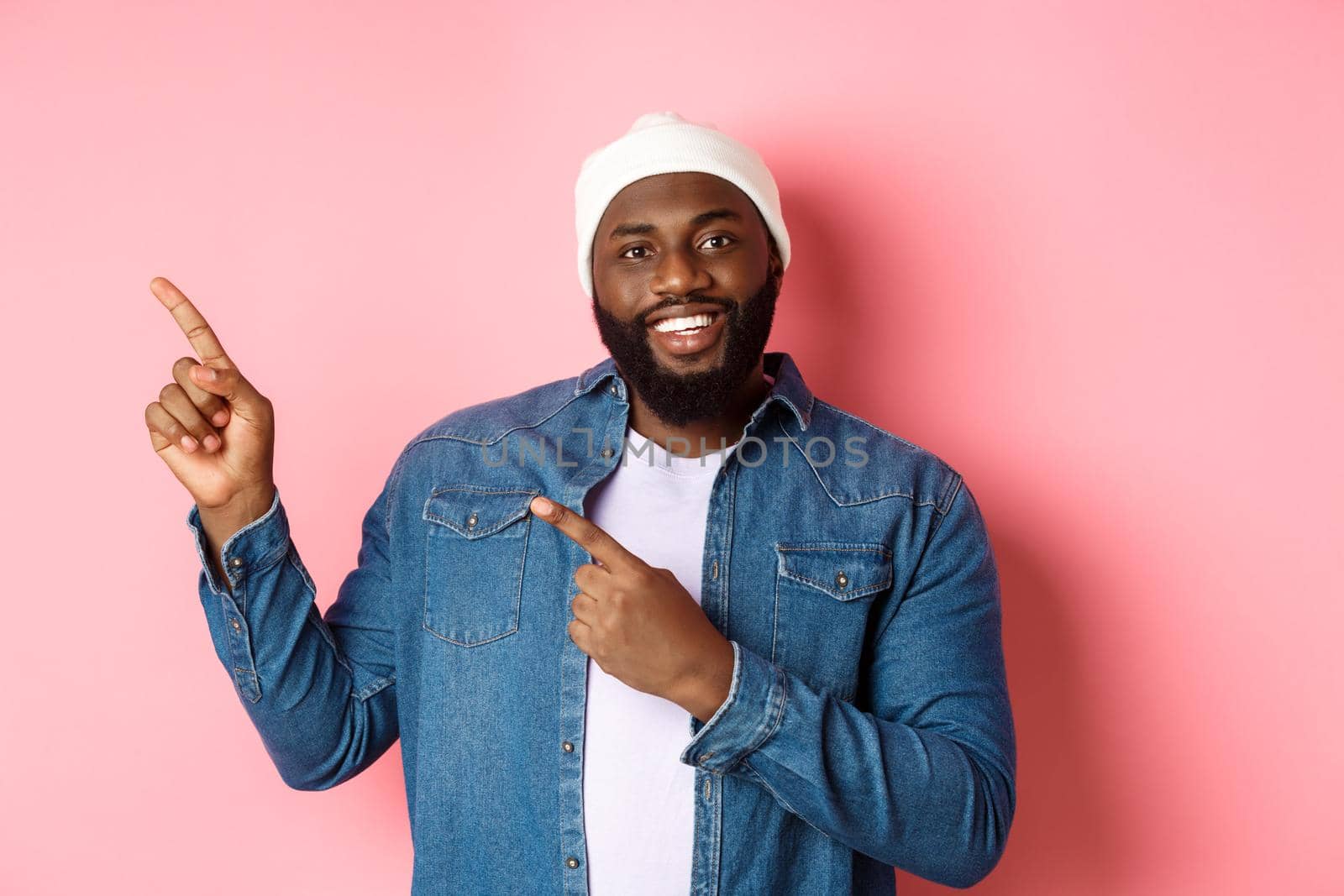 Happy african american man pointing fingers upper left corner, showing promo offer logo, smiling pleased, wearing beanie with denim jacket, pink background.