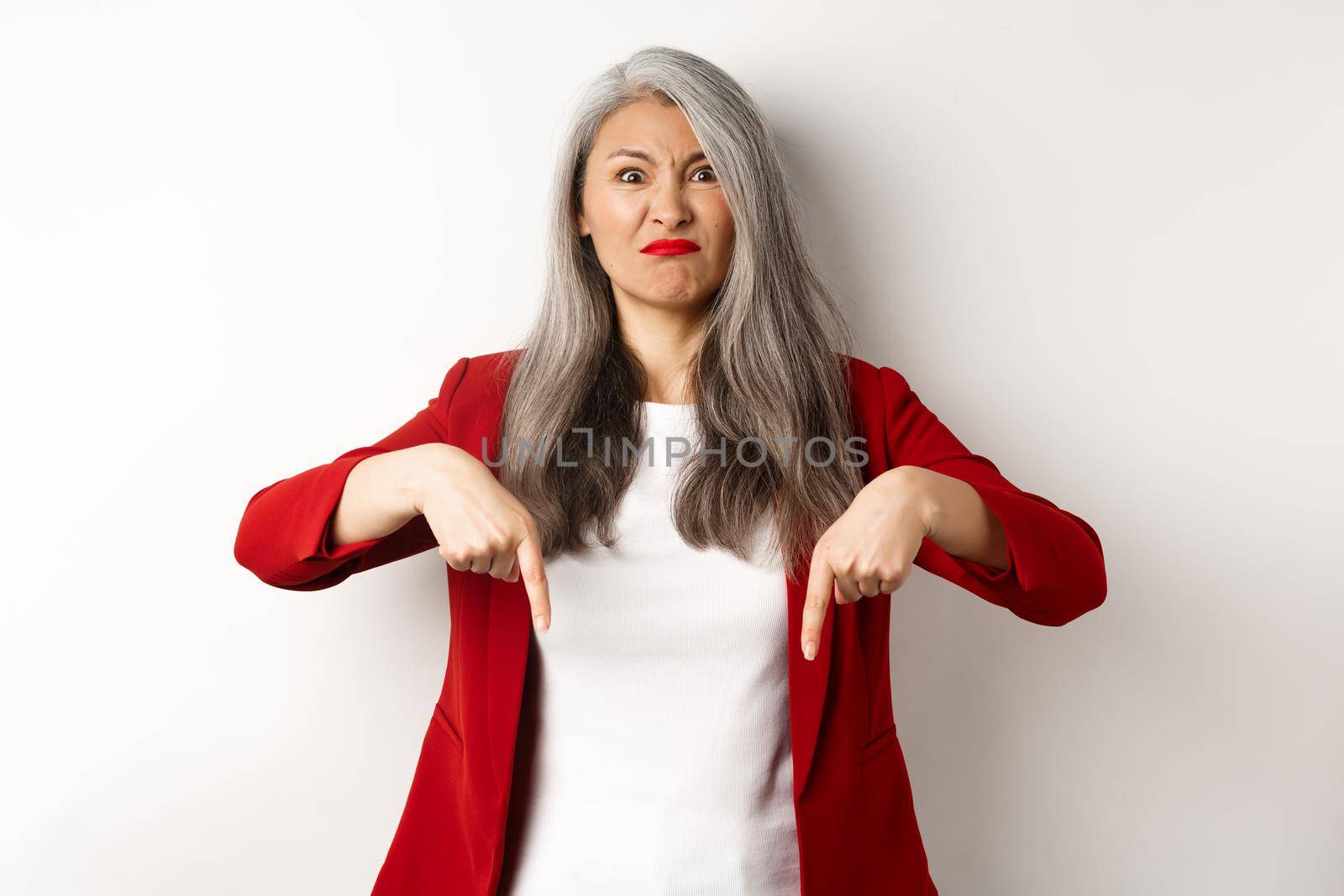 Disappointed and angry asian employer pointing fingers down, looking displeased and upset, standing over white background.