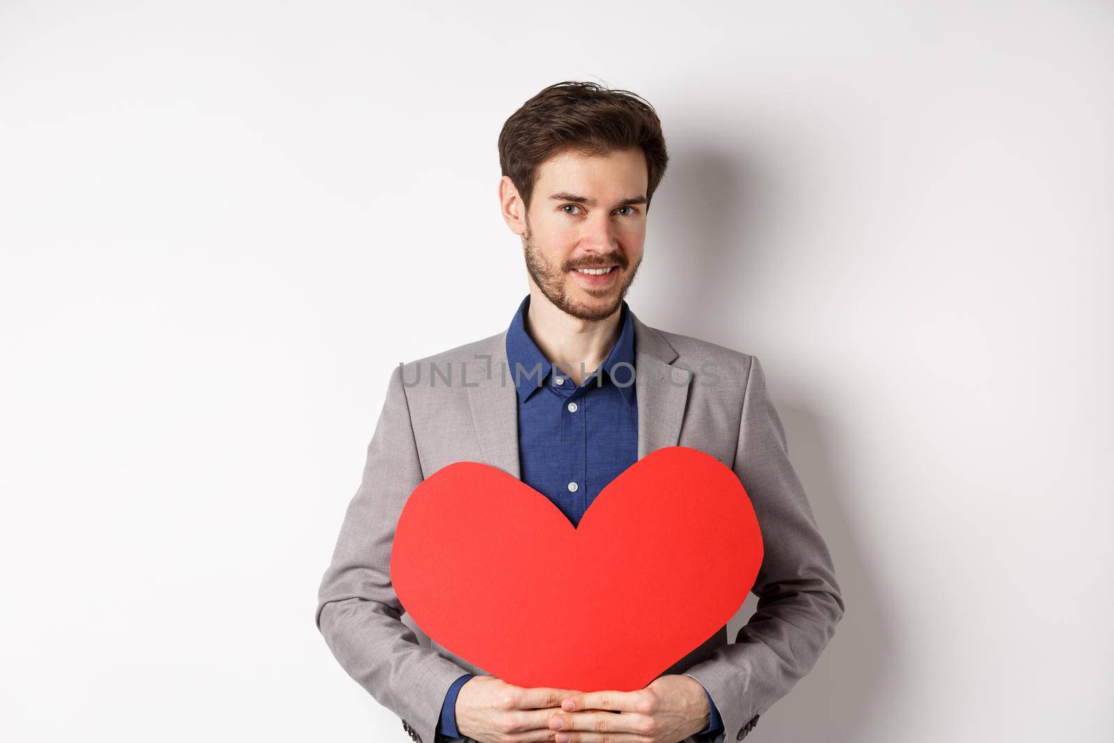 Handsome bearded man wishing happy valentines day, holding red heart cutout and smiling, going on romantic date in fancy suit, standing over white background.