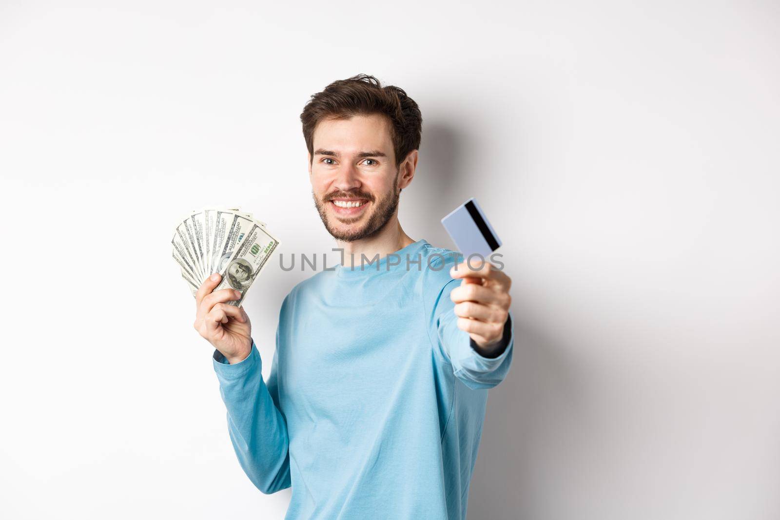 Smiling caucasian man holding money and giving you plastic credit card, standing on white background by Benzoix