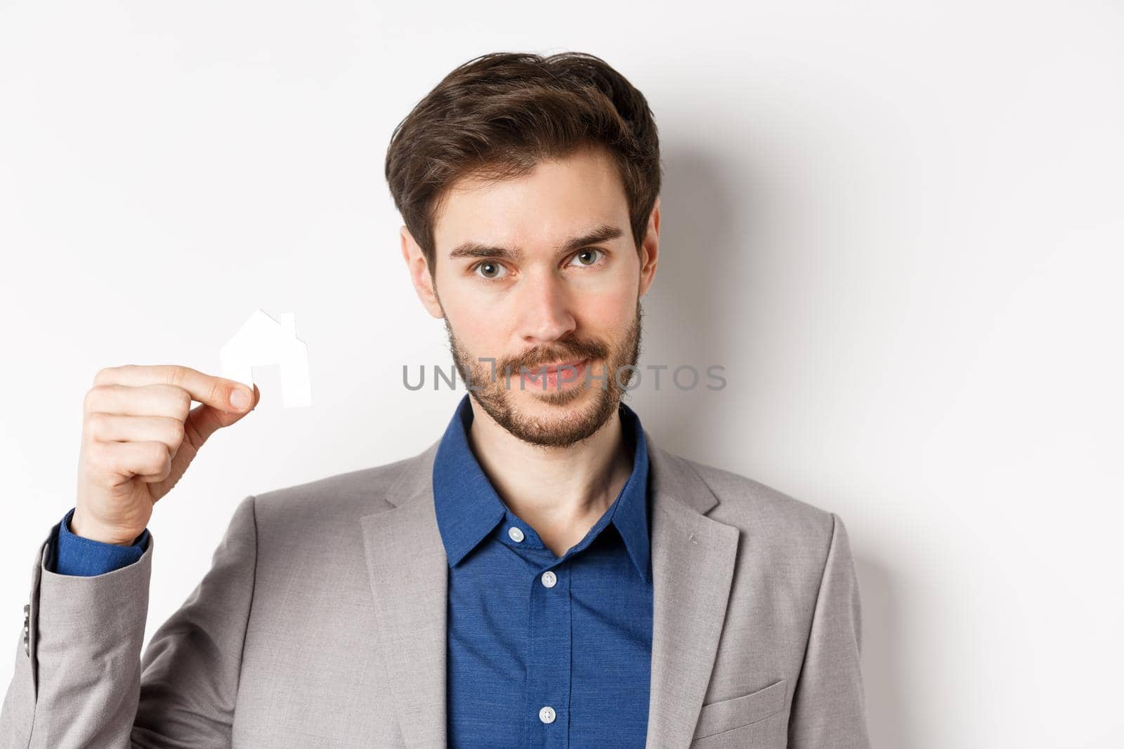 Real estate and insurance concept. Close up portrait of handsome bearded man in suit showing small paper house cutout and look assertive, white background.