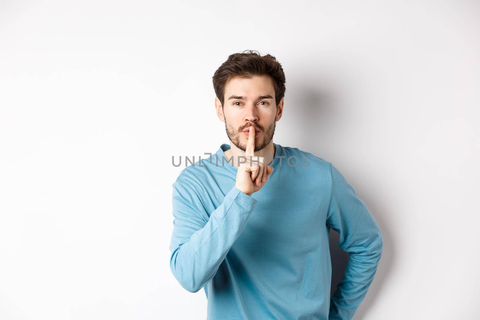 Attractive bearded man asking to keep quiet, showing taboo hush gesture and looking at camera calm, standing over white background.