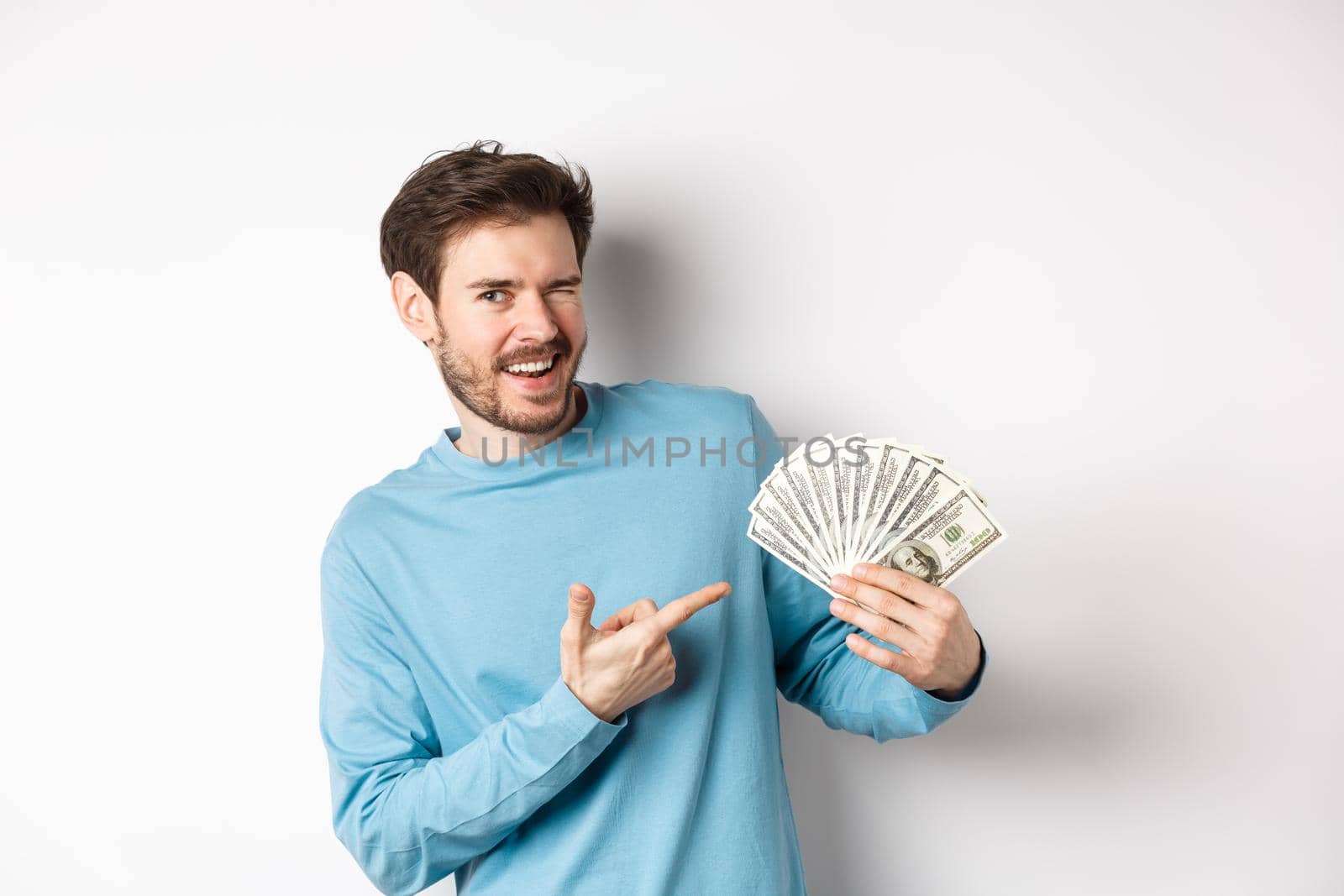 Handsome rich guy winking at camera, showing income, pointing finger at earned money and smiling, standing over white background by Benzoix