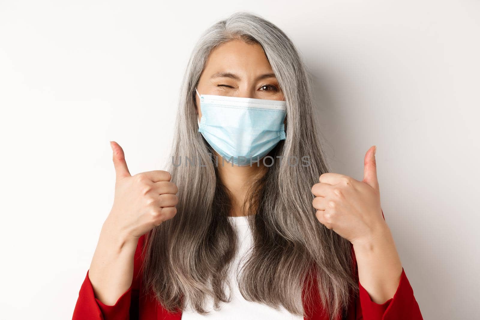 Covid, pandemic and business concept. Cheerful asian businesswoman in medical mask, winking and showing thumbs-up in approval, white background.
