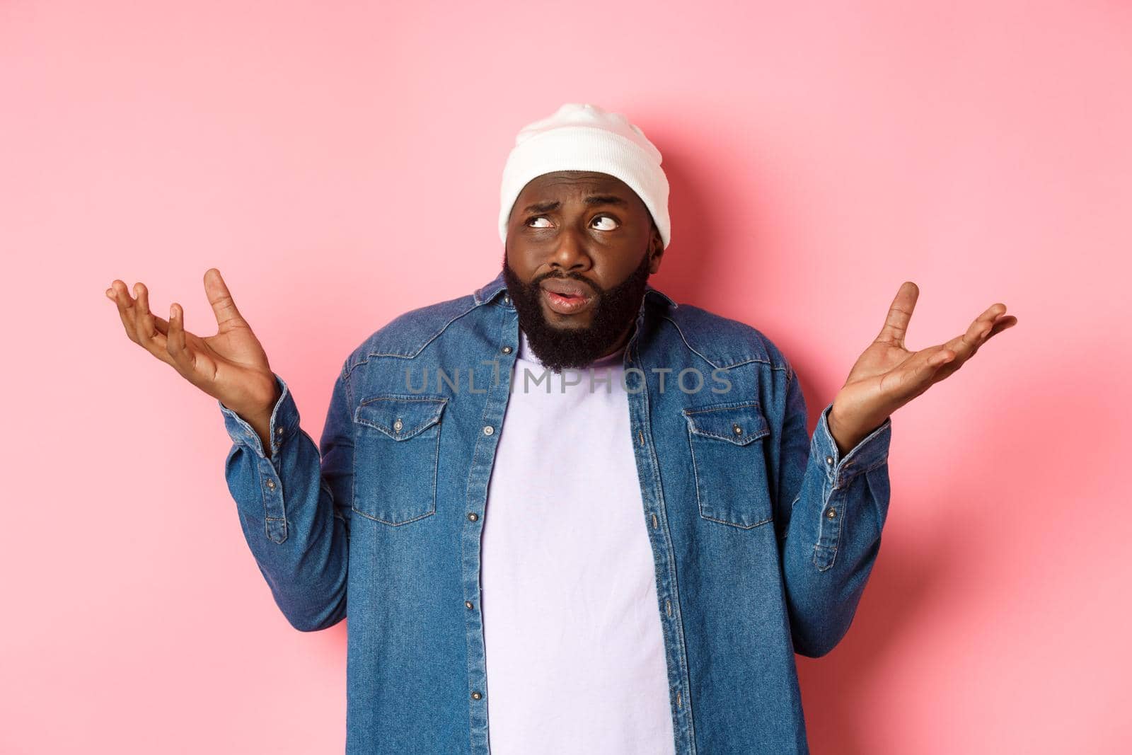 Confused african-american man in beanie looking at upper left corner doubtful, shrugging uncertain, standing over pink background.