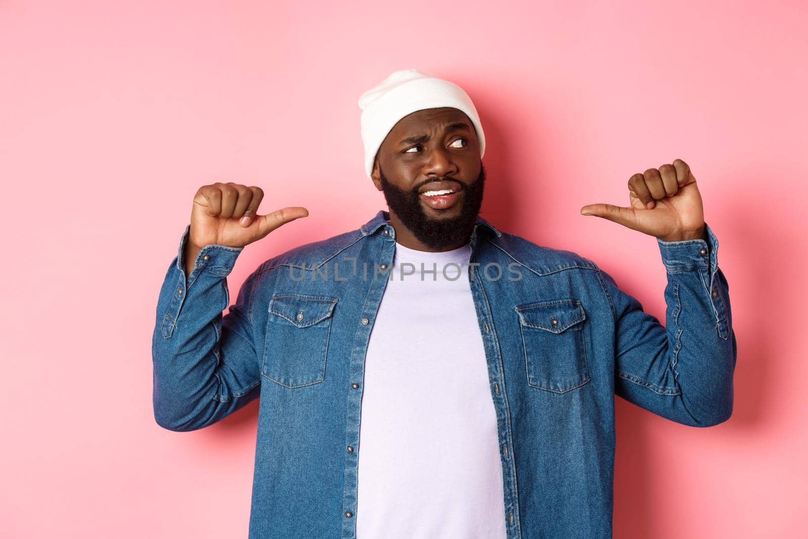 Confused Black man staring right and pointing at himself unsure, having doubts, standing over pink background.