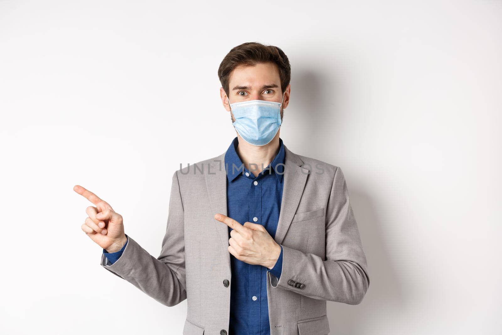 Smiling businessman in suit and medical mask showing advertisement, pointing fingers left and looking friendly, white background. Covid-19 and pandemic concept.