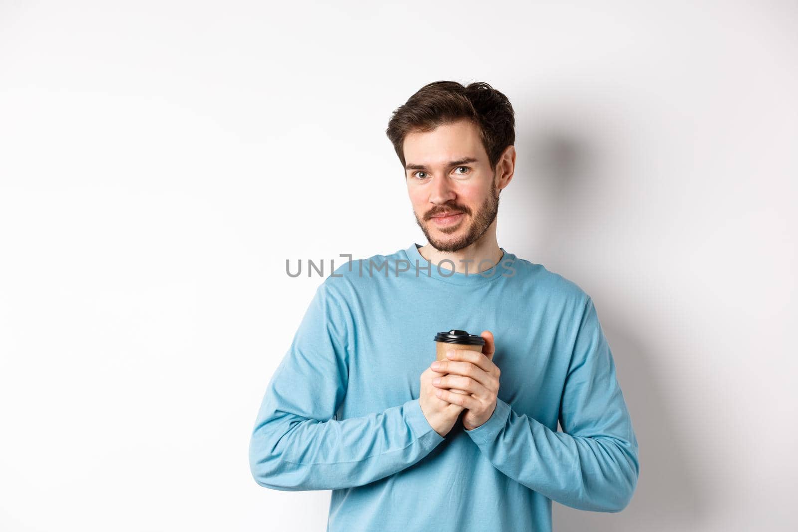 Handsome caucasian man warm-up hands with cup of coffee, smiling at camera and looking tenderly, standing on white background.