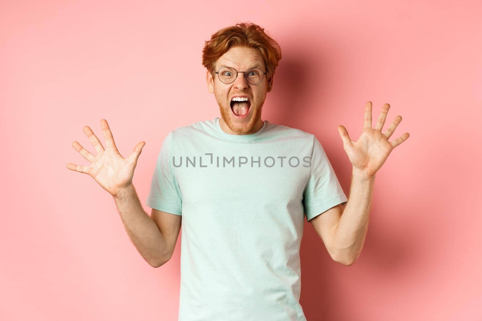 Angry and pressured young man losing temper, spread hands sideways and screaming with furious face, standing in glasses and t-shirt against pink background by Benzoix