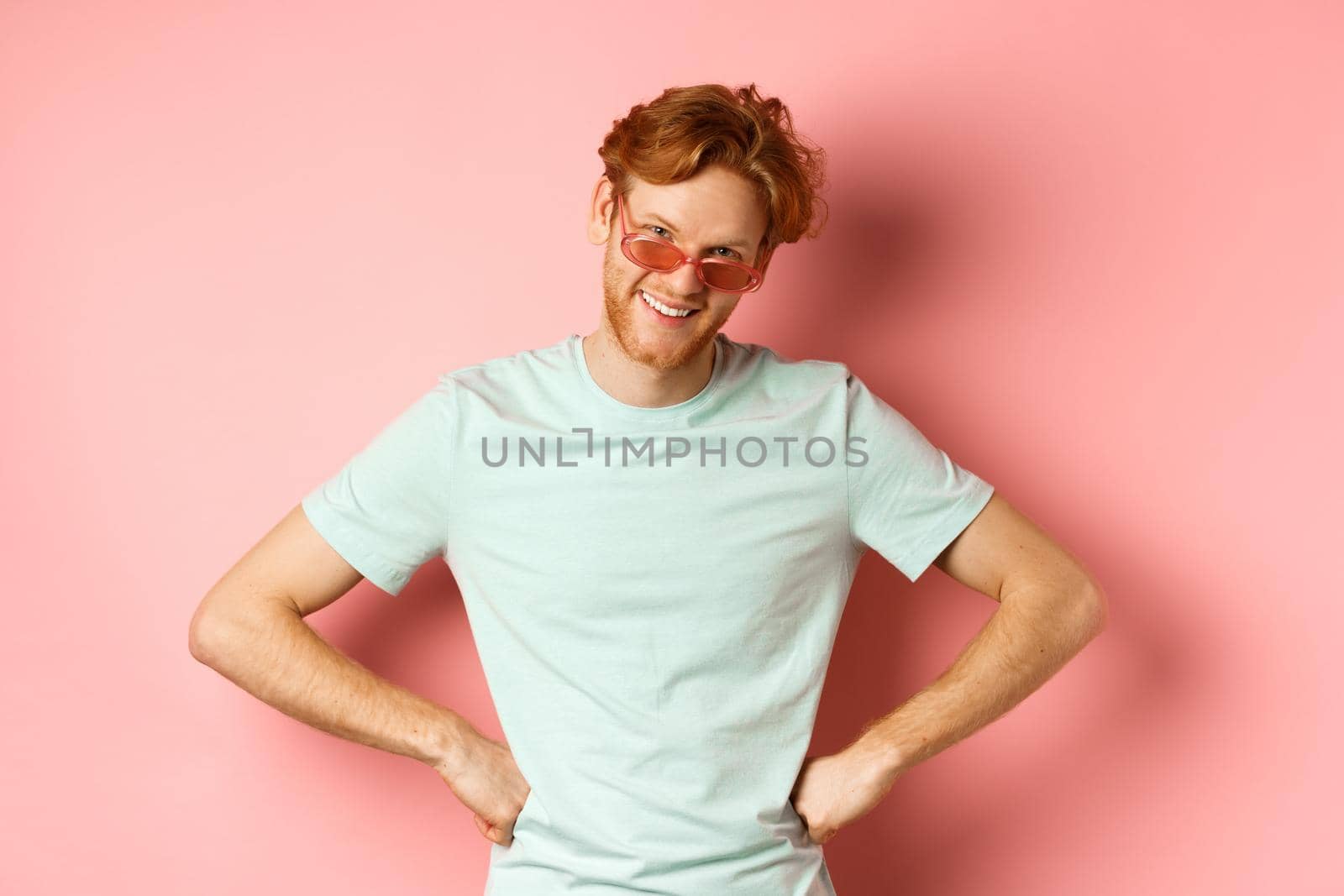 Tourism and vacation concept. Funny bearded man with red hair, wearing glasses, smiling and looking from under forehead with hands on hips, standing over pink background by Benzoix