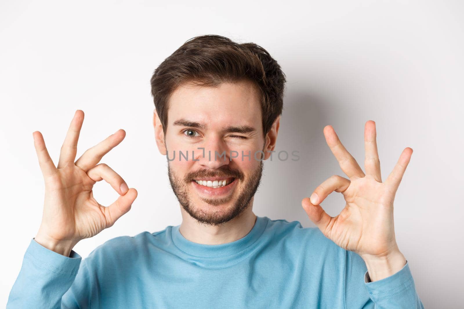 Close up of handsome bearded man winking and smiling, showing okay signs, assure all good, standing on white background.
