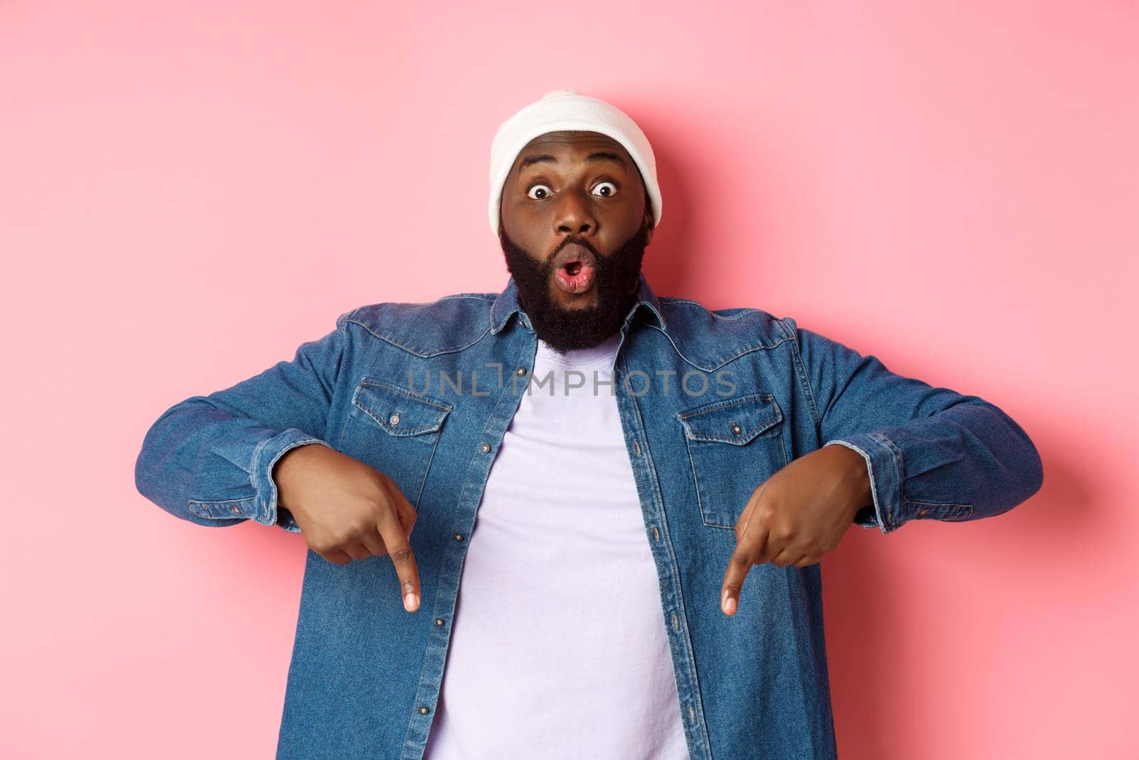 Amazed and impressed african-american guy showing advertisement, staring at camera happy and pointing fingers down, pink background by Benzoix