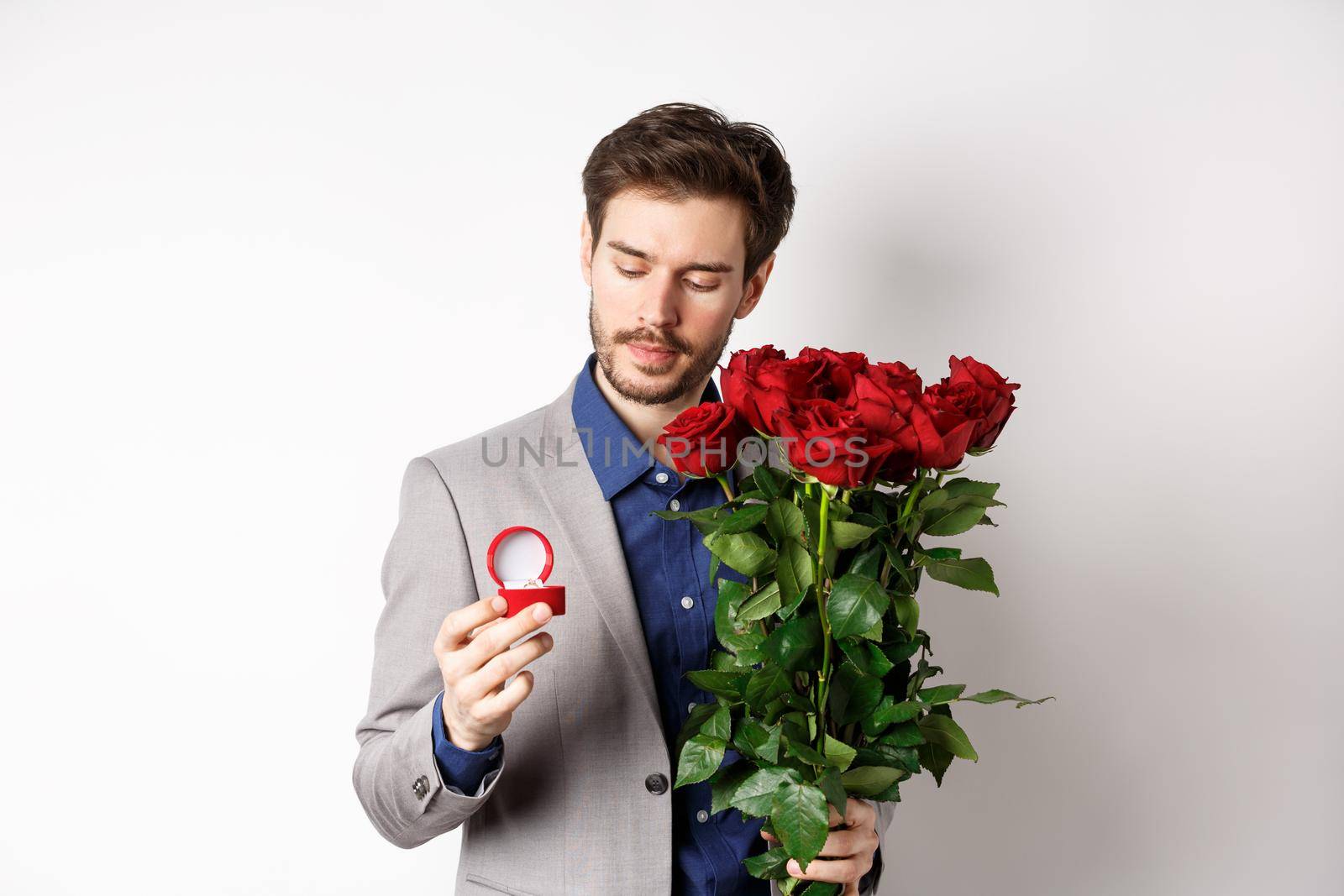 Romantic man in suit looking pensive at engagement ring, going to make a marriage proposal on Valentines day, holding bouquet of roses, standing over white background by Benzoix