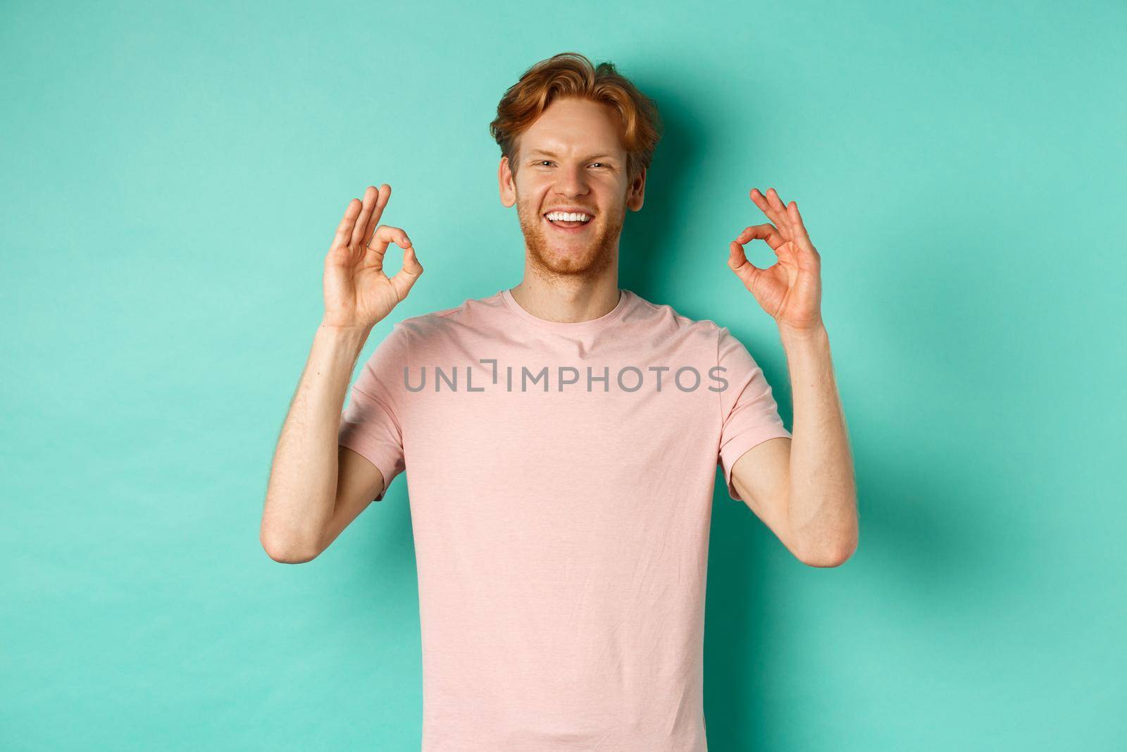 Attractive young man in t-shirt smiling satisfied, nod in approval and showing OK sign, approve and agree with something cool, standing over turquoise background.