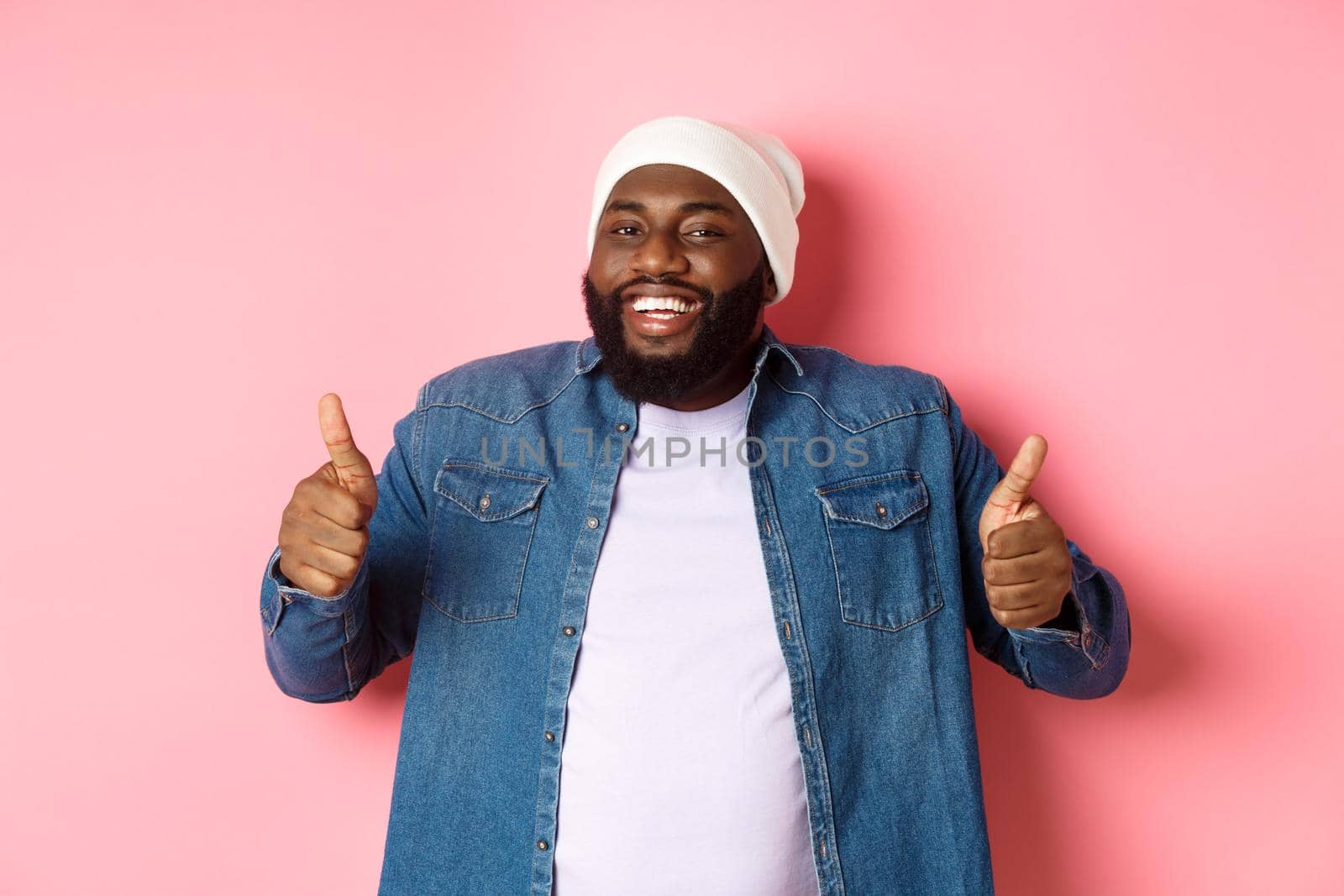 Satisfied handsome black man looking happy, showing thumbs-up and smiling, give approve and praise product, recommending company, pink background.