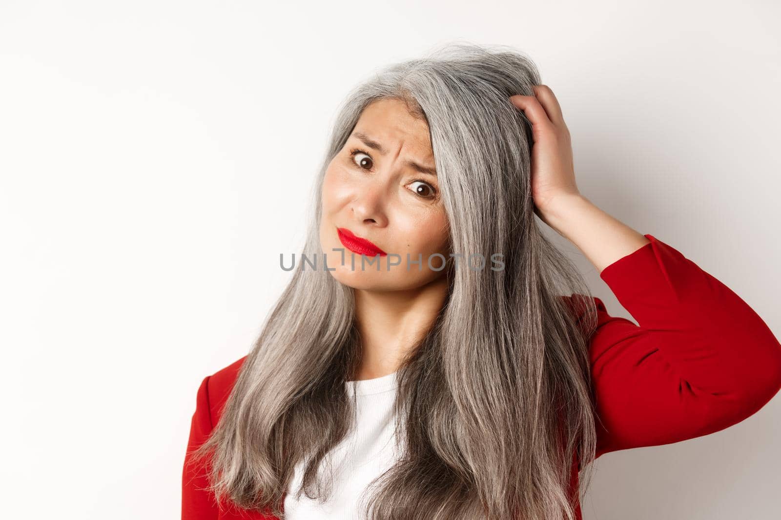 Close up portrait of confused asian female manager scratching head and looking questioned at camera, standing over white background by Benzoix