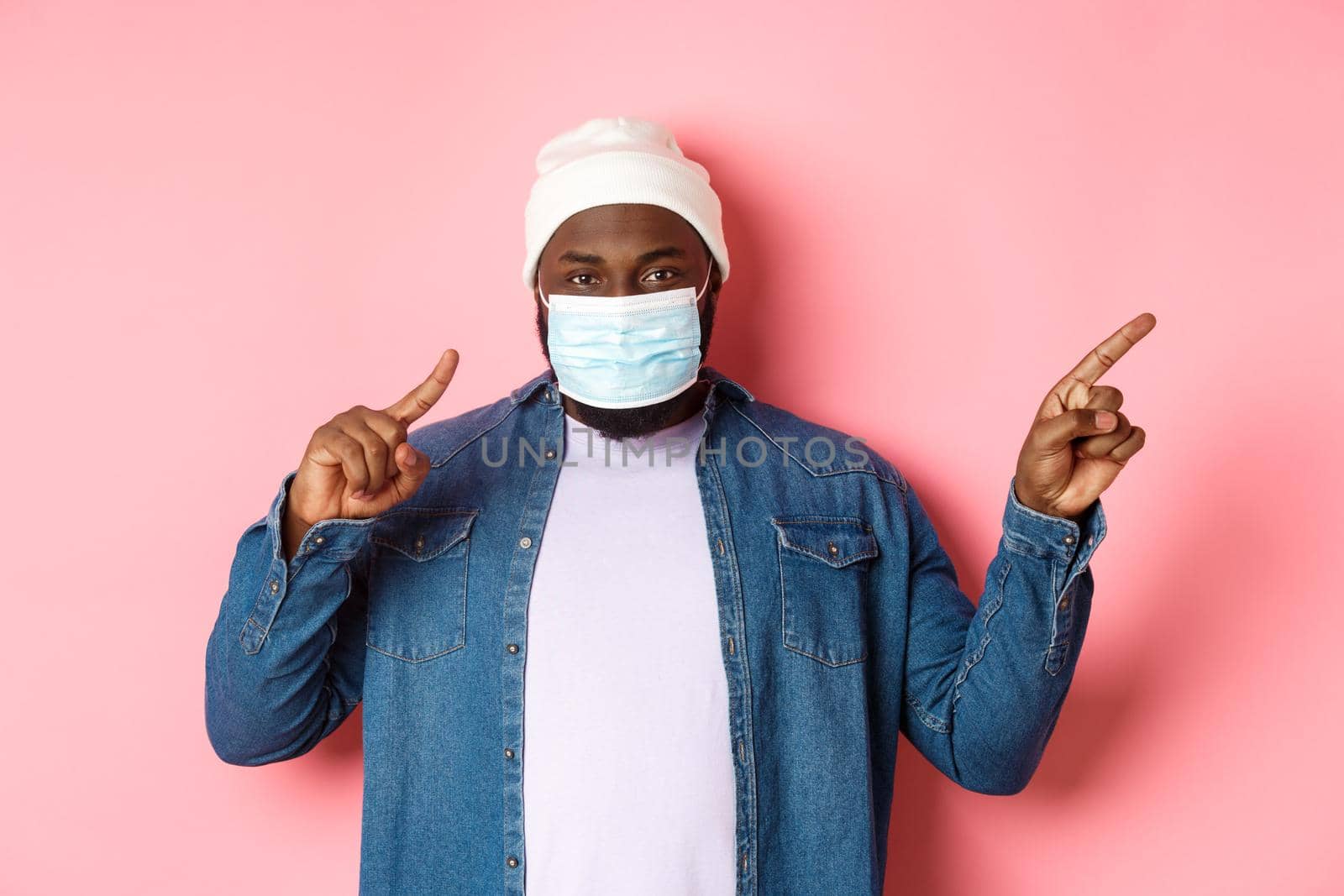 Covid-19, lifestyle and lockdown concept. Smiling african-american man in beanie and face mask pointing fingers right, showing copy space, standing over pink background.
