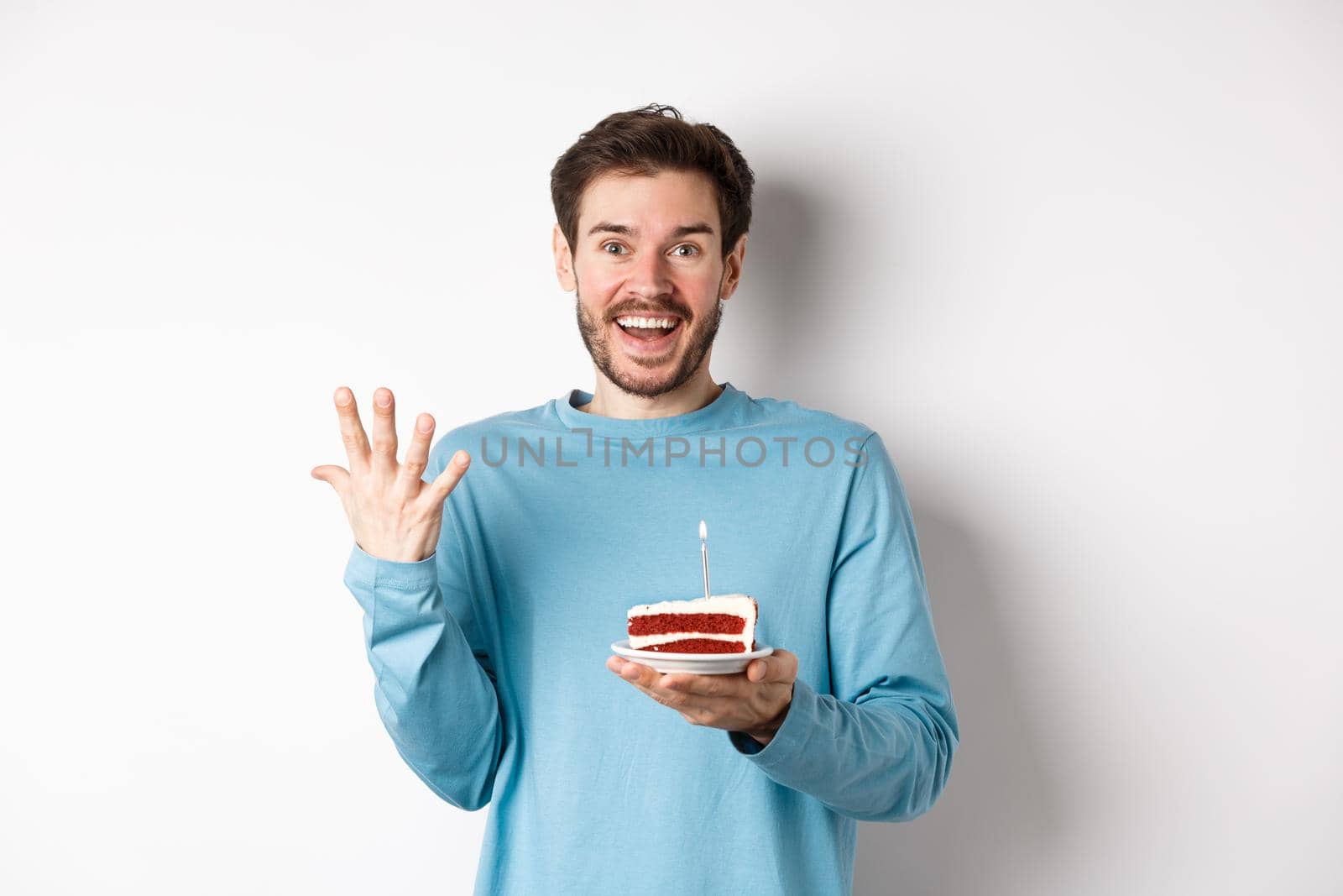 Excited man receive birthday surprise, holding bday cake and smiling happy, standing over white background, making wish on lit candle by Benzoix