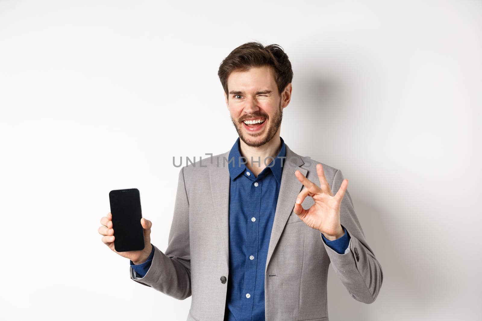 E-commerce and online shopping concept. Successful businessman making money on smartphone, showing empty phone screen and okay sign, winking happy at camera.