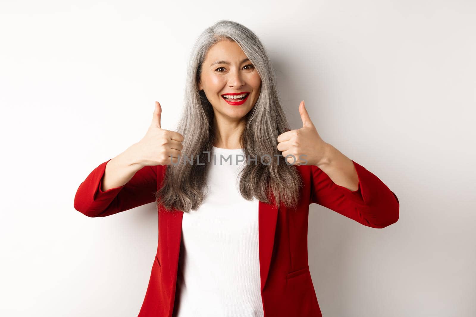 Portrait of senior asian woman showing thumb-up in approval, wearing red blazer for office work, recommend company, standing over white background by Benzoix