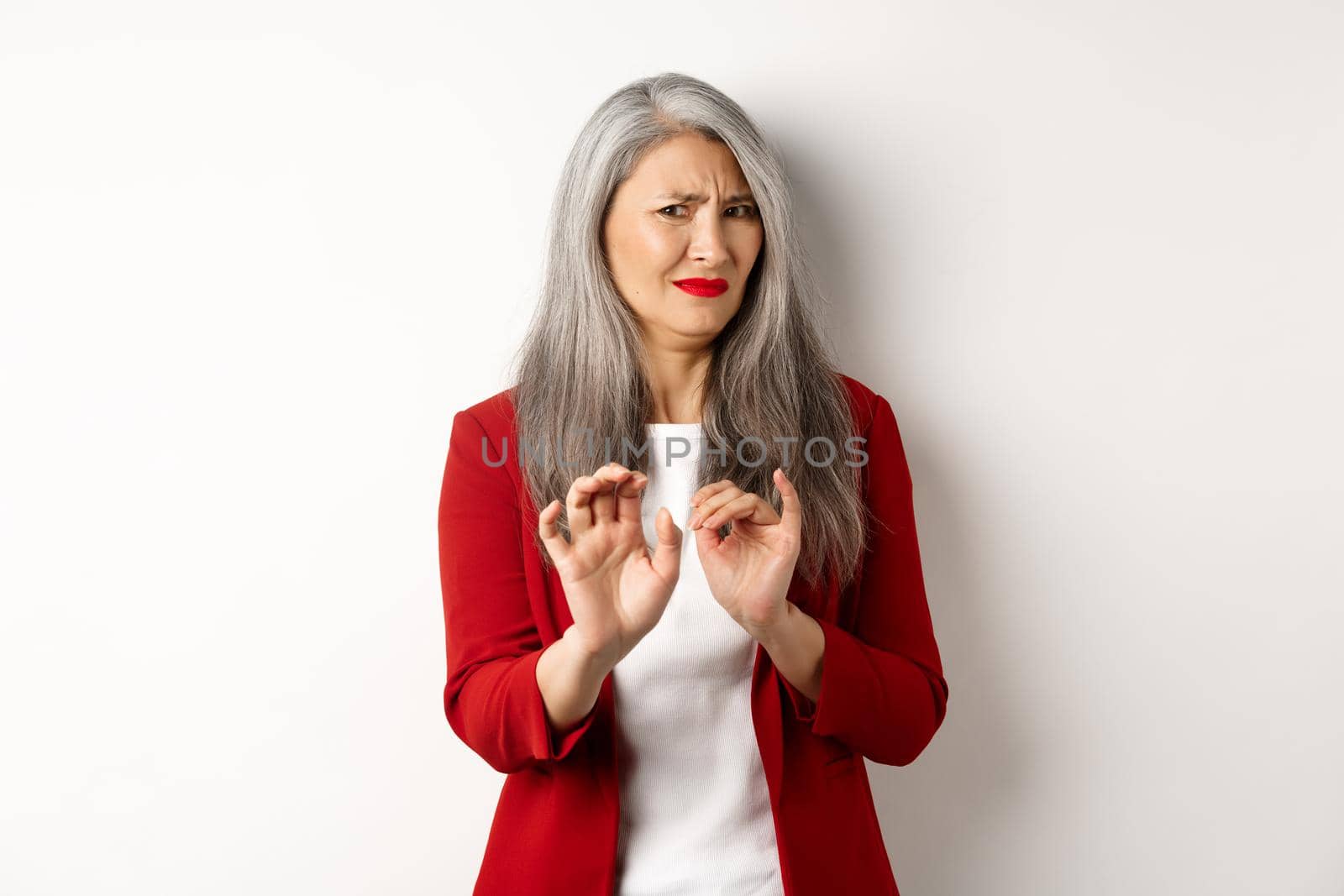 Disgusted asian businesswoman with grey hair, wearing red blazer and makeup, rejecting something disgusting, showing stop sign, white background.