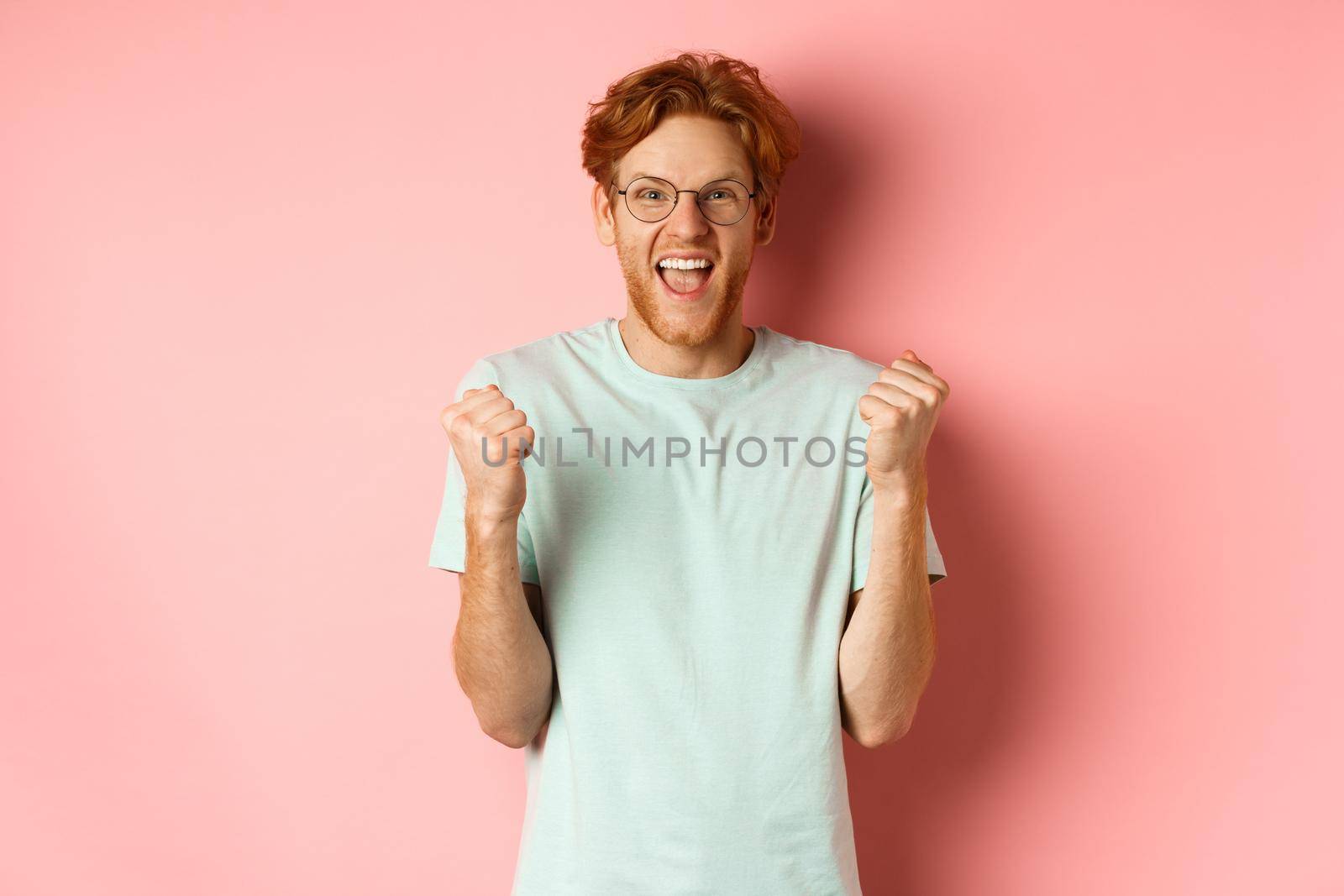 Excited young man winning prize, shouting with joy and triumph, making fist pump and saying yes, standing over pink background by Benzoix