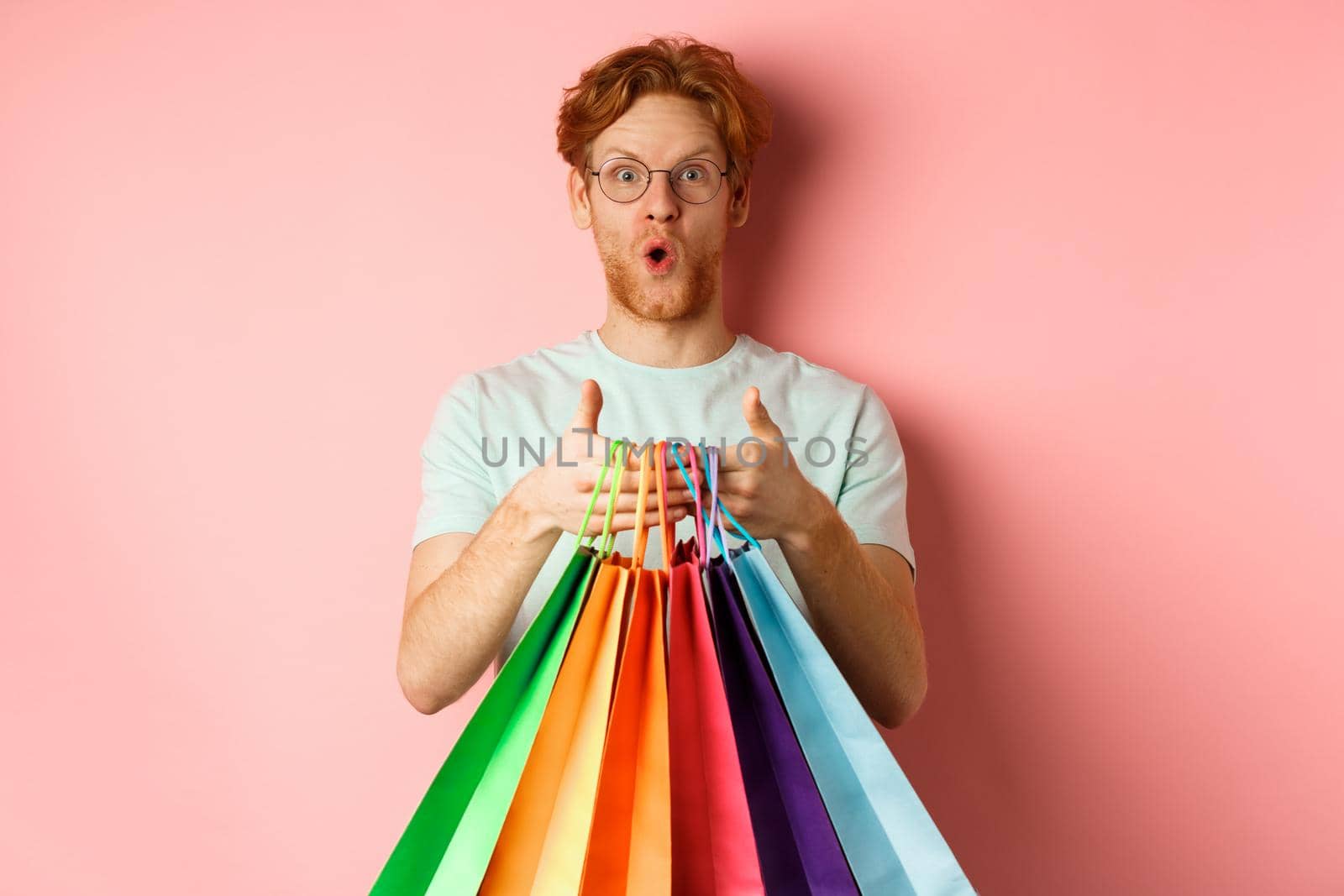 Young handsome man amazed with special discounts, buying gifts, holding shopping bags and standing over pink background.