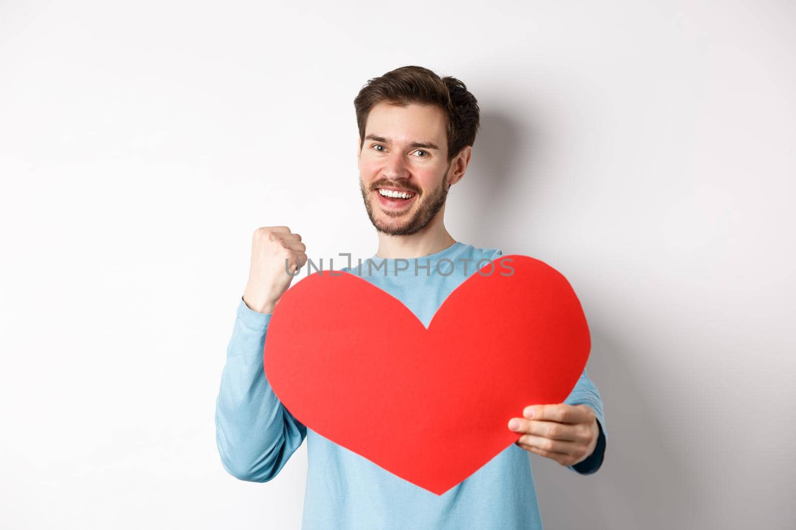 Valentines day. Happy boyfriend triumphing, saying yes and showing valentine red heart, smiling as winning girls love, standing over white background.