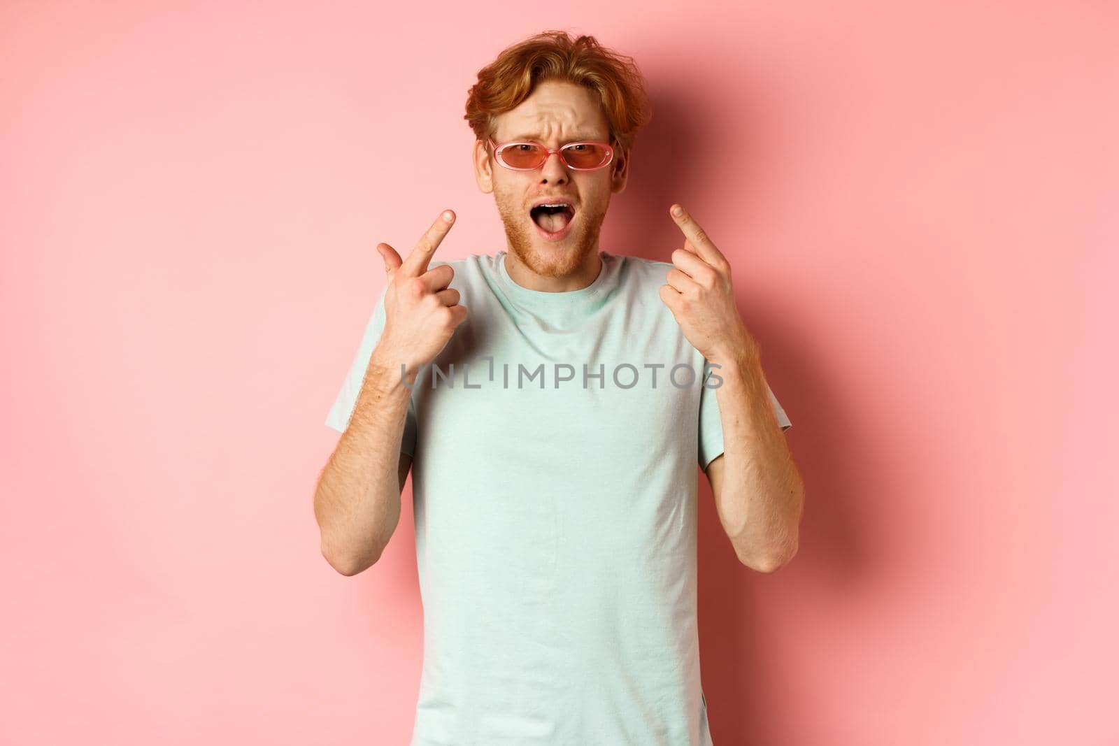 Young handsome man with red hair and beard pointing fingers at sunglasses, showing new glasses, standing over pink background by Benzoix