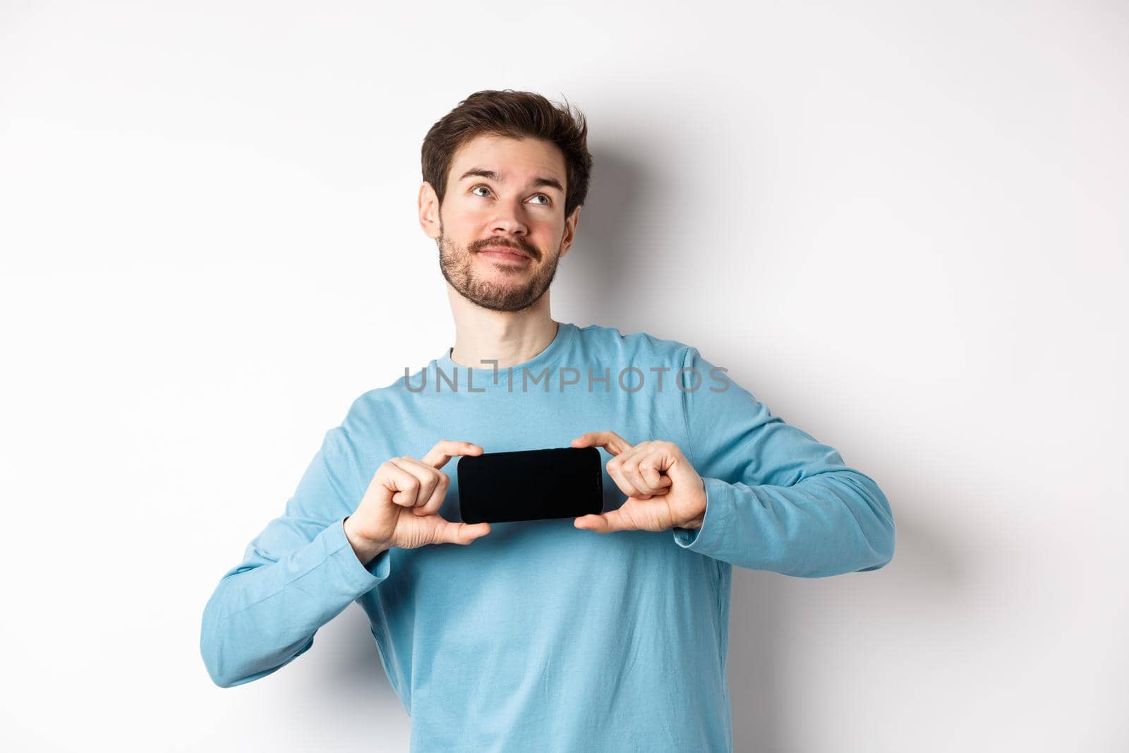 E-commerce and shopping concept. Smiling young man showing mobile screen and dreaming about something, looking at upper right corner nostalgic, standing over white background.