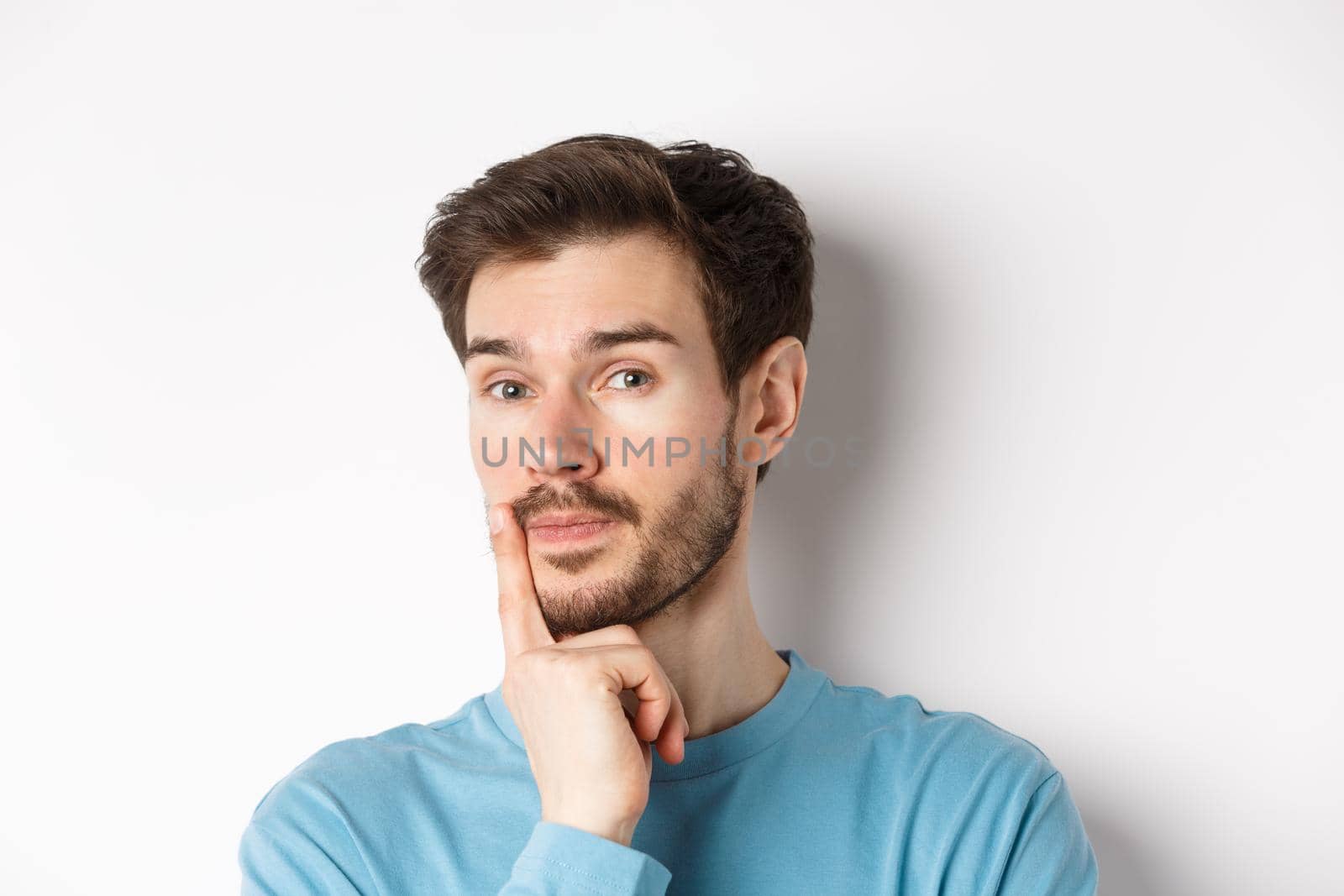 Image of thoughtful young man with beard, making choice, touching lip and looking pensive at camera, deciding something, white background.