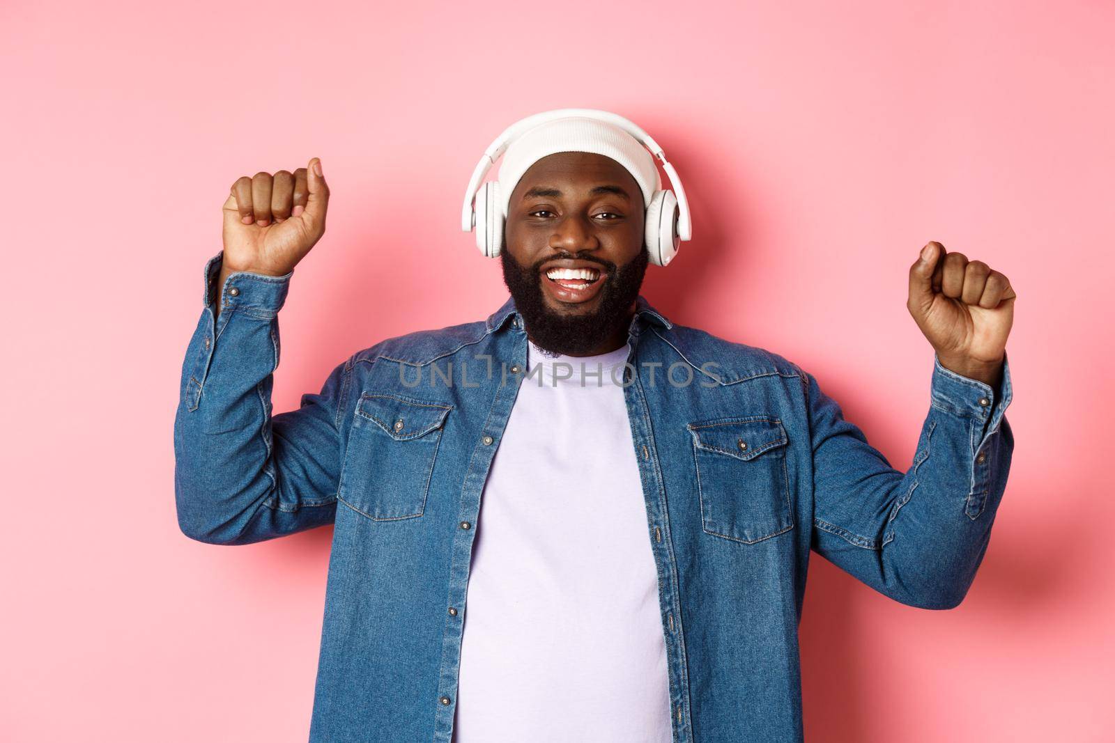 Happy young african-american man dancing and listening music in headphones, pump fists up and smiling, standing over pink background.