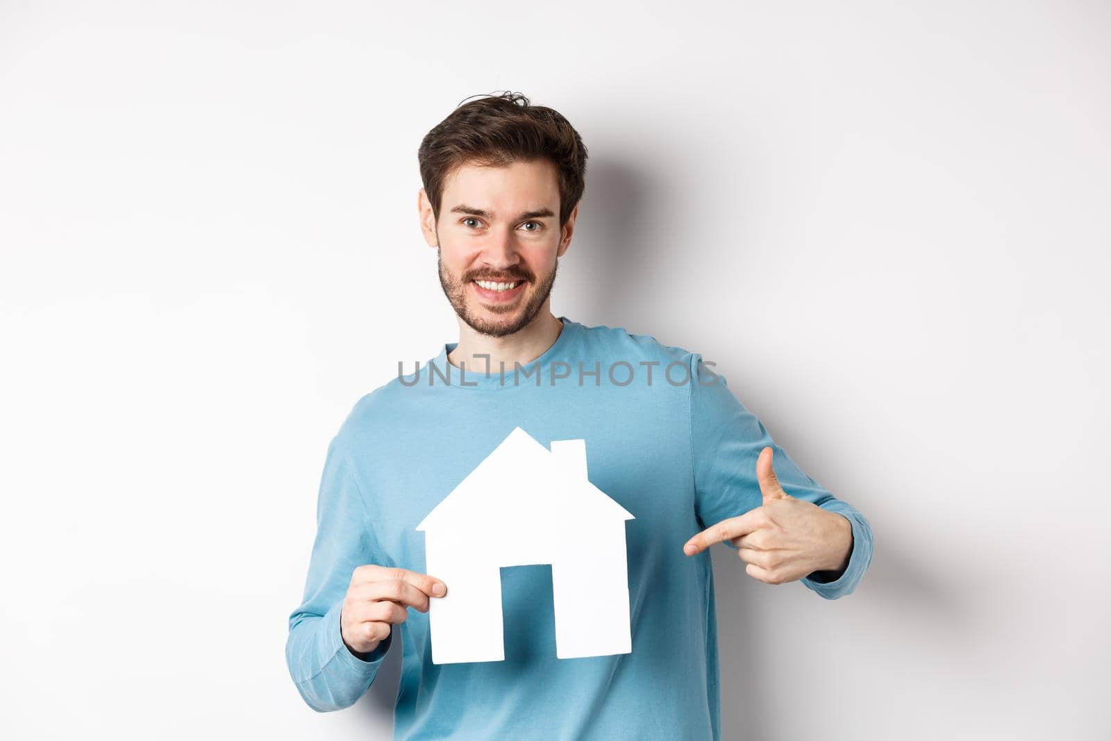 Real estate and insurance concept. Smiling young man pointing at paper house cutout and looking happy, buy apartment, standing over white background.