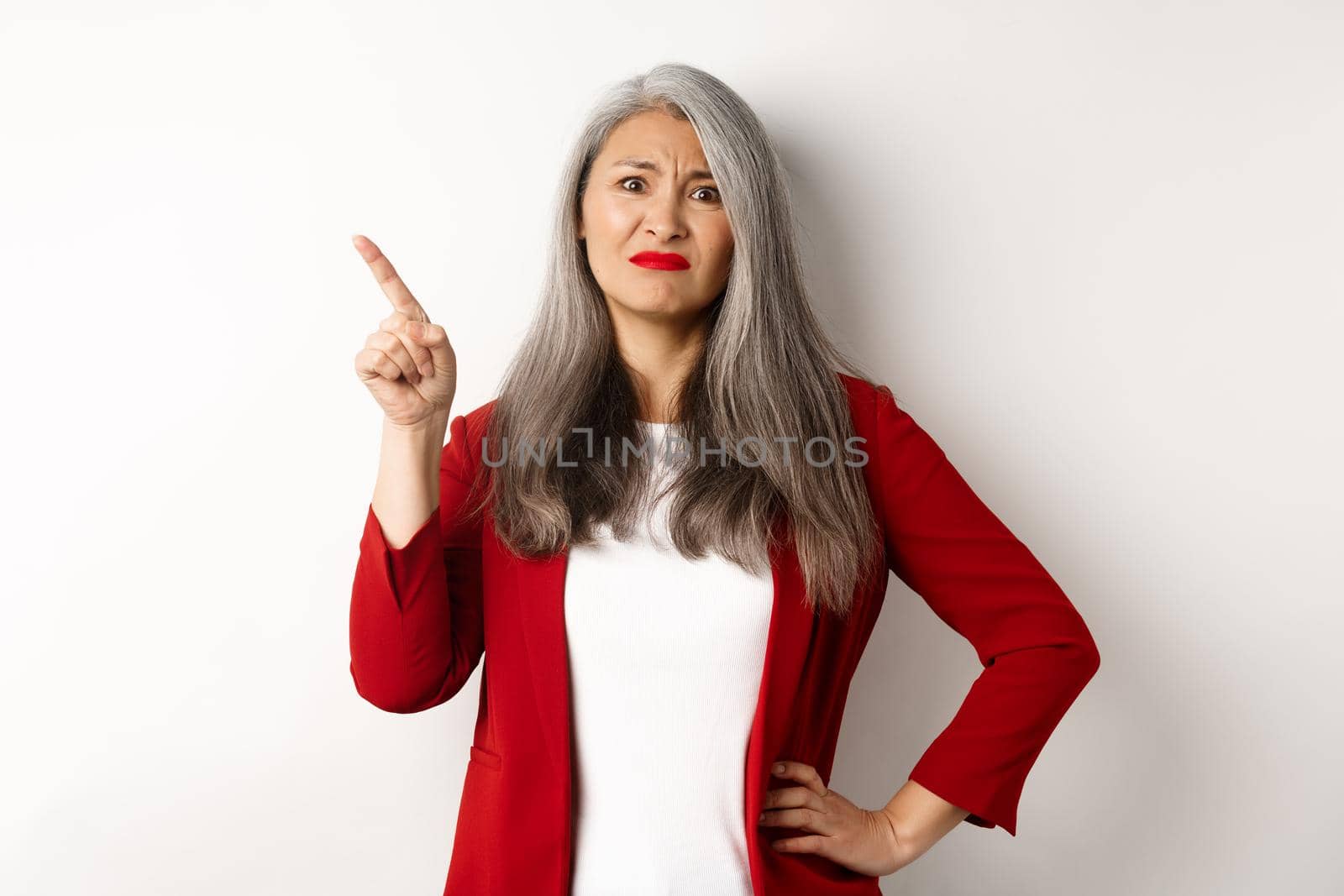 Skeptical asian businesswoman in red blazer grimacing, pointing upper left corner and looking dissatisfied, disapprove something, standing over white background by Benzoix