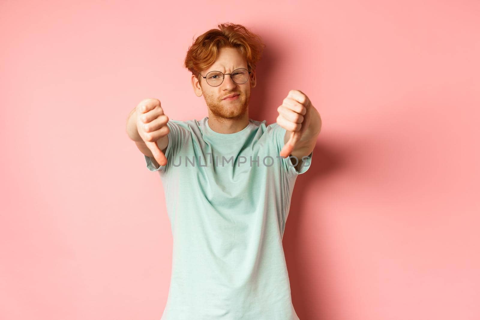 Disappointed young man in glasses, with messy red haircut, showing thumbs down and grimacing unsatisfied, express dislike, standing over pink background.