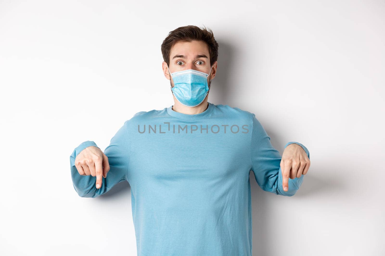 Covid-19, health and quarantine concept. Amazed man checking out promo logo, pointing fingers down, wearing medical mask from coronavirus, white background by Benzoix