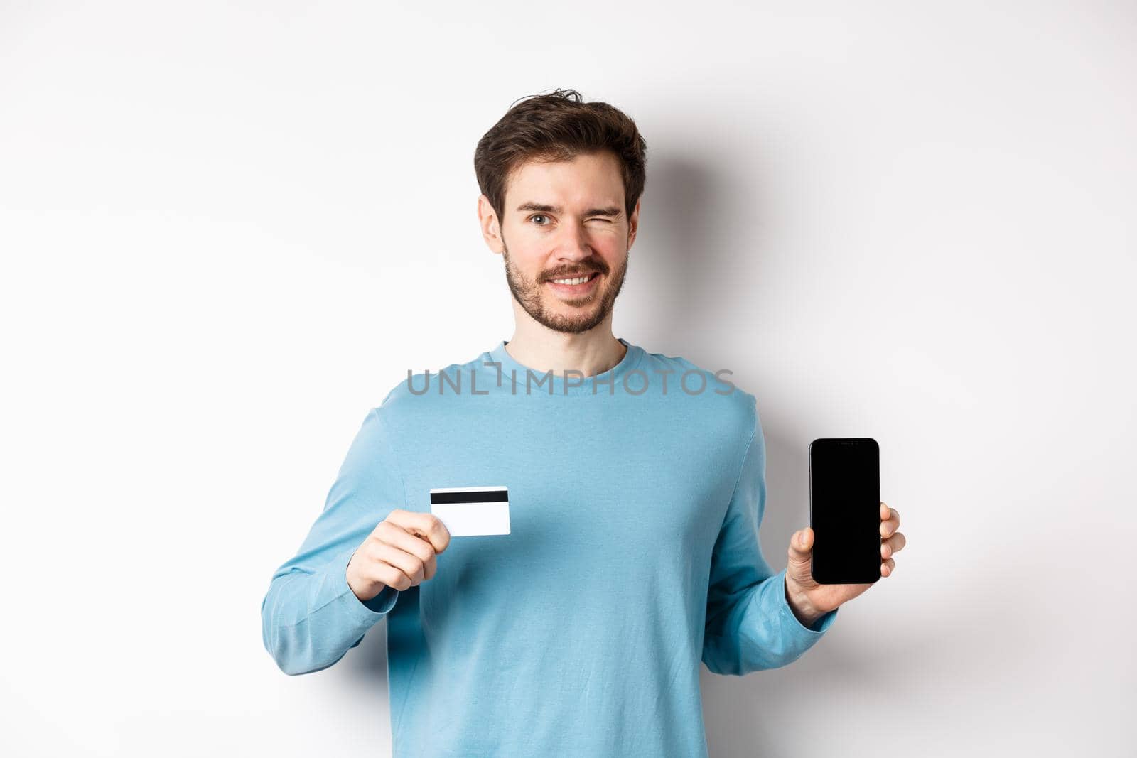 Young man in casual shirt showing empty smartphone screen and plastic credit card, winking and smiling at camera, standing on white background.
