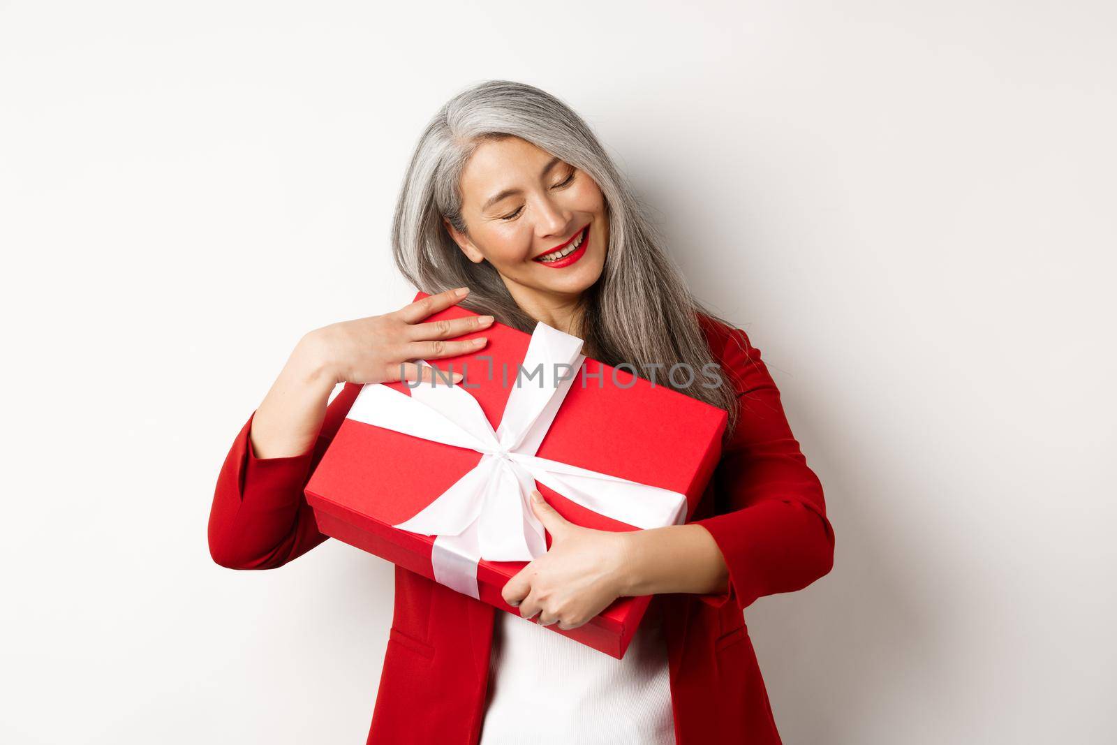 Valentine day concept. Beautiful asian senior woman hugging box with gift, smiling and looking happy, receiving present on holiday, white background.