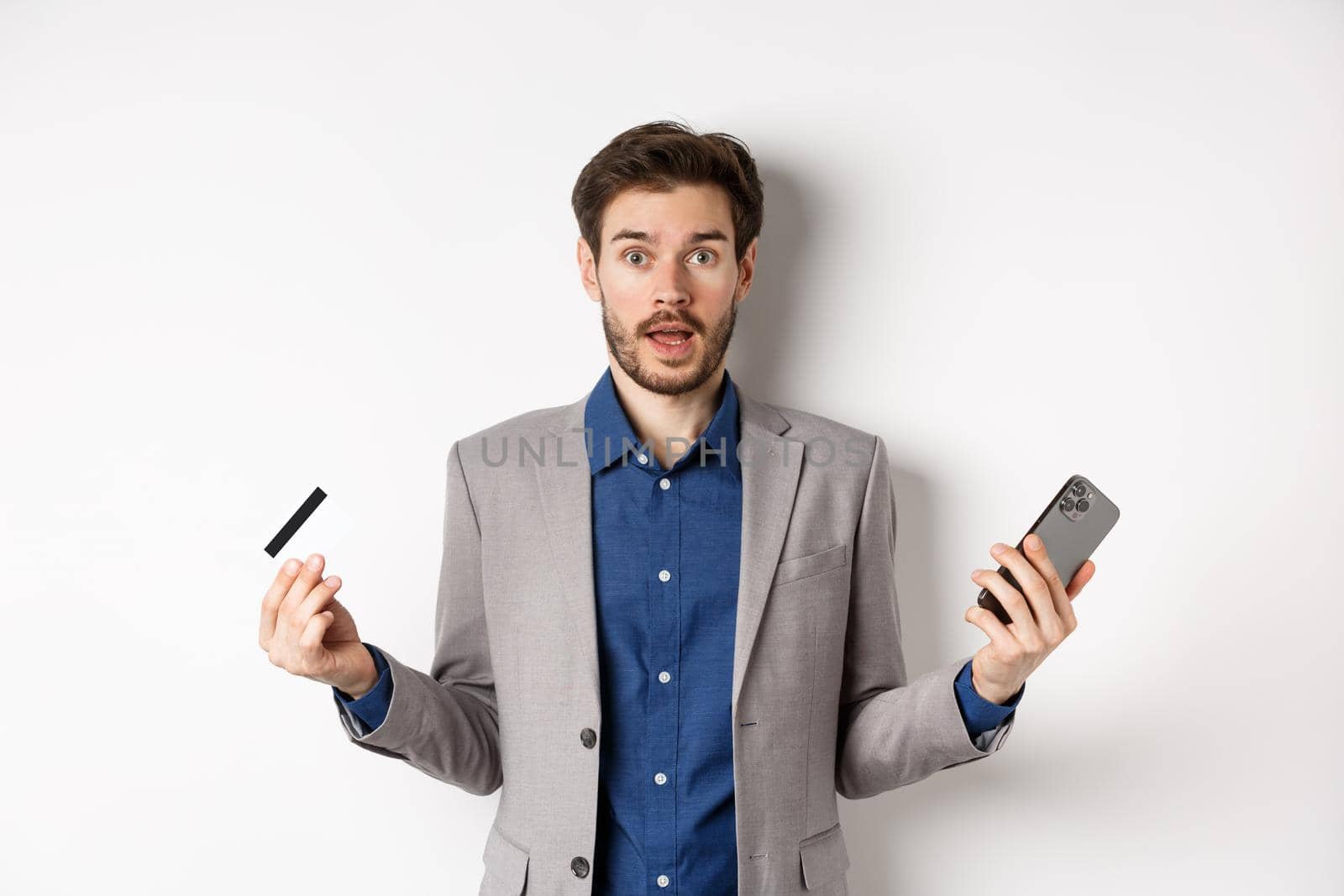 Online shopping. Confused man in suit spread hands sideways, holding plastic credit card with mobile phone and shrugging cueless, standing on white background by Benzoix