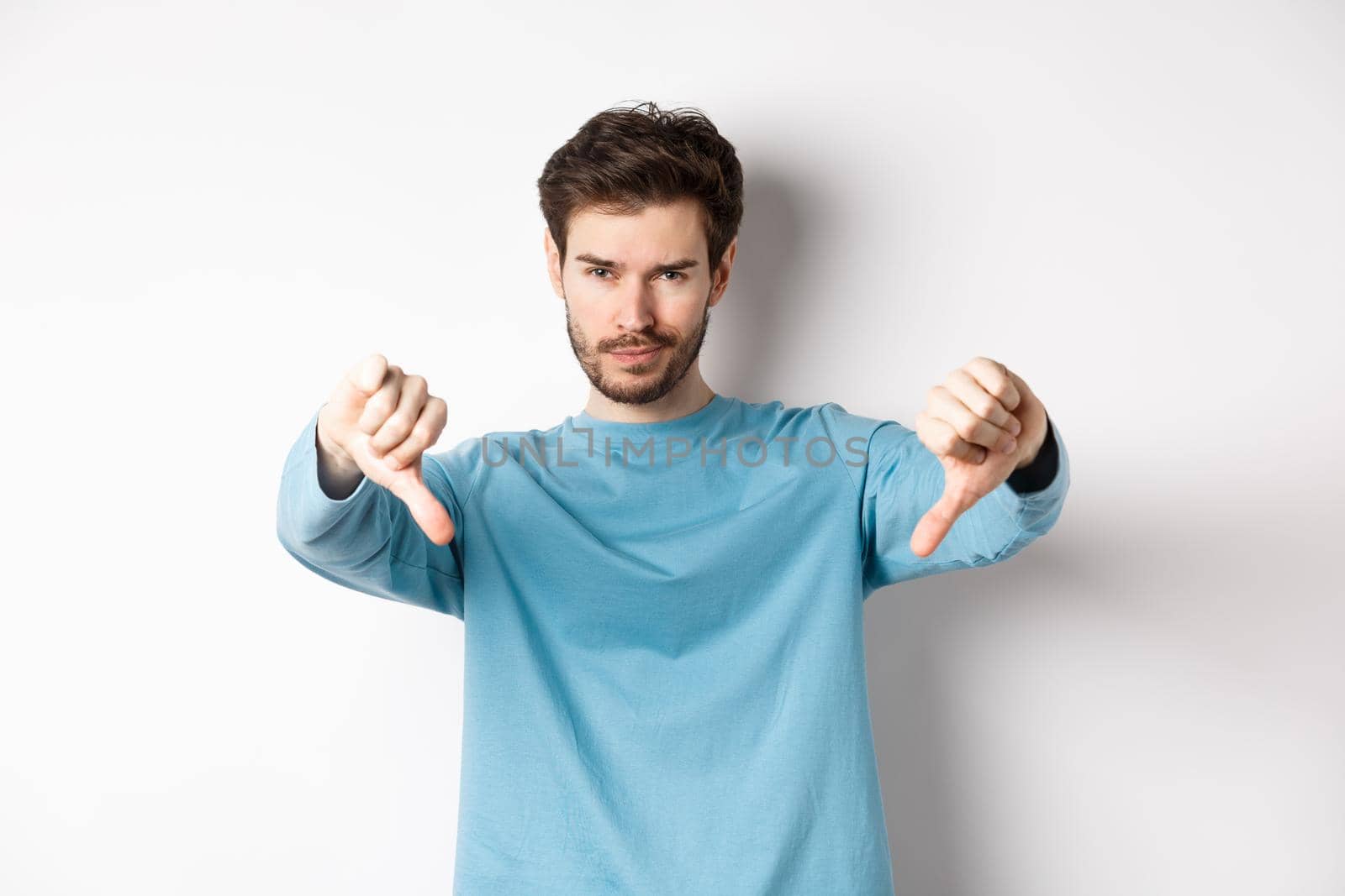 Skeptical handsome guy with beard, showing thumbs down in disapproval, dislike something bad, standing over white background by Benzoix