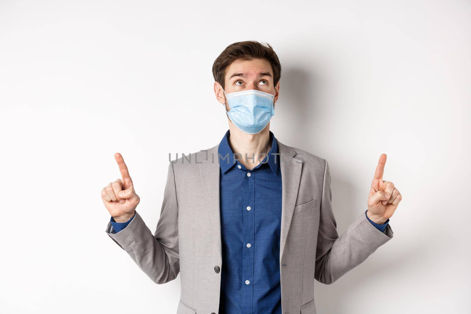 Covid-19, pandemic and business concept. Handsome businessman in medical mask and suit, looking and pointing up with dreamy face, white background.