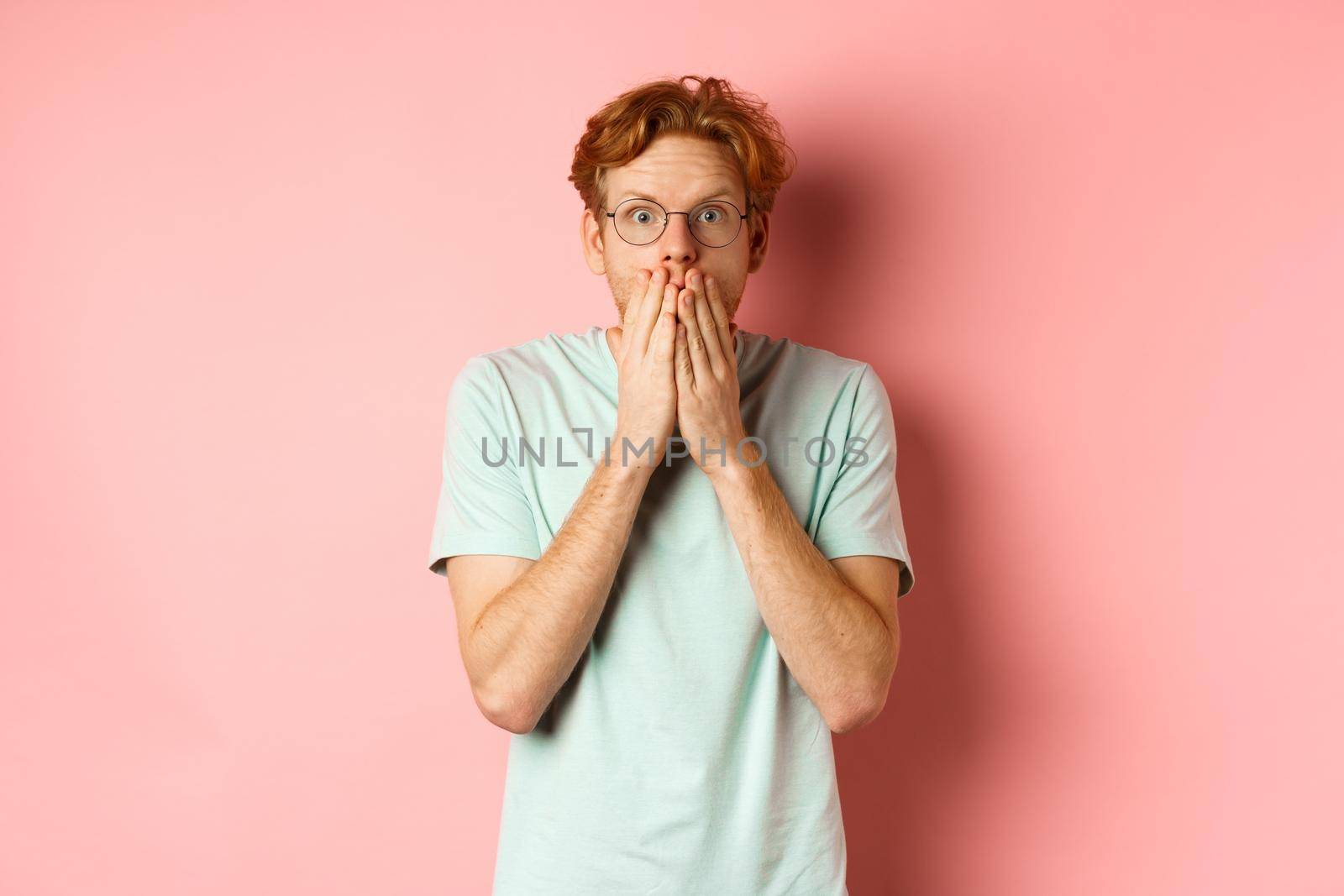 Shocked and speechless redhead guy covering mouth, staring at camera startled, standing in t-shirt against pink background by Benzoix